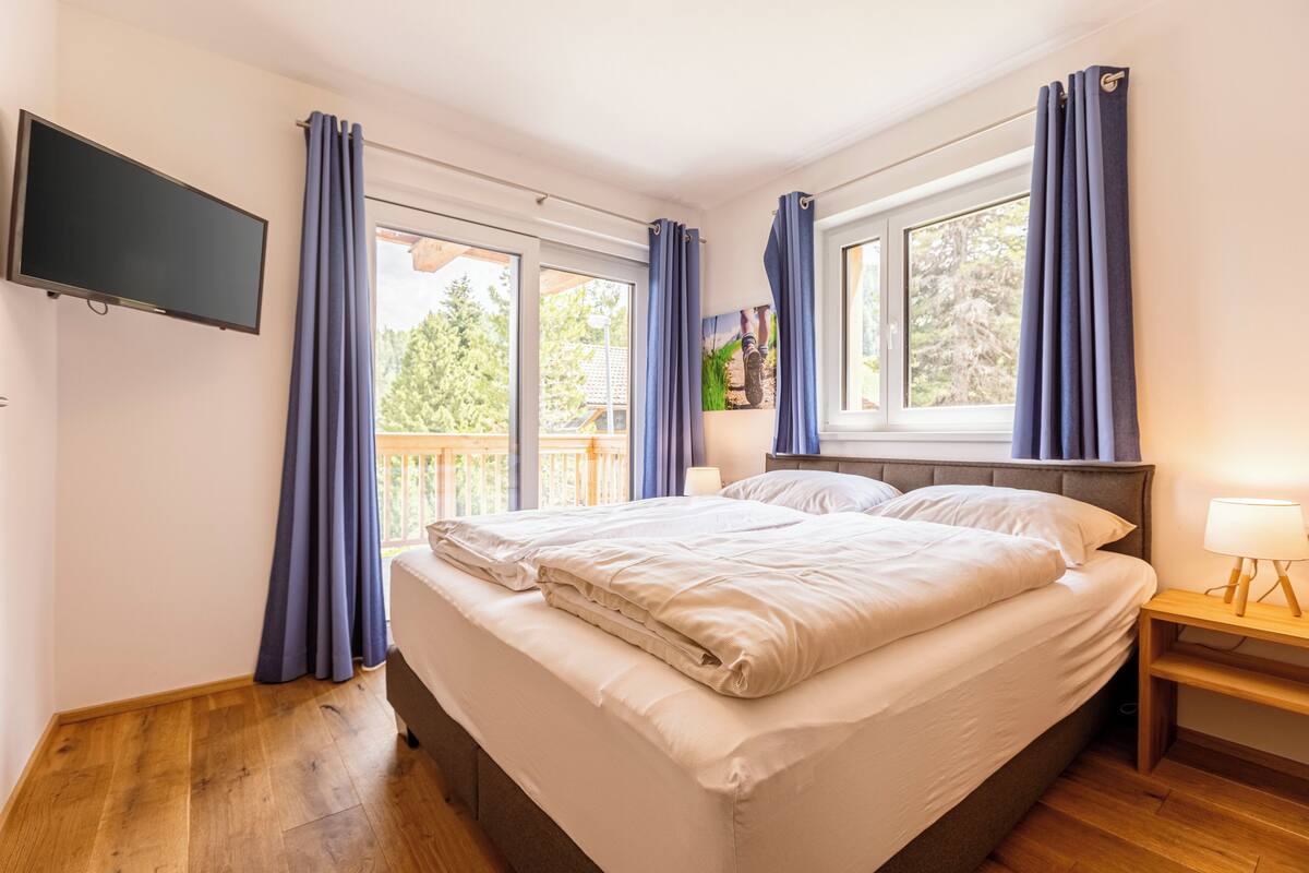 A bedroom with a bed, a TV mounted on the wall, a nightstand with a lamp, and a window with curtains.
