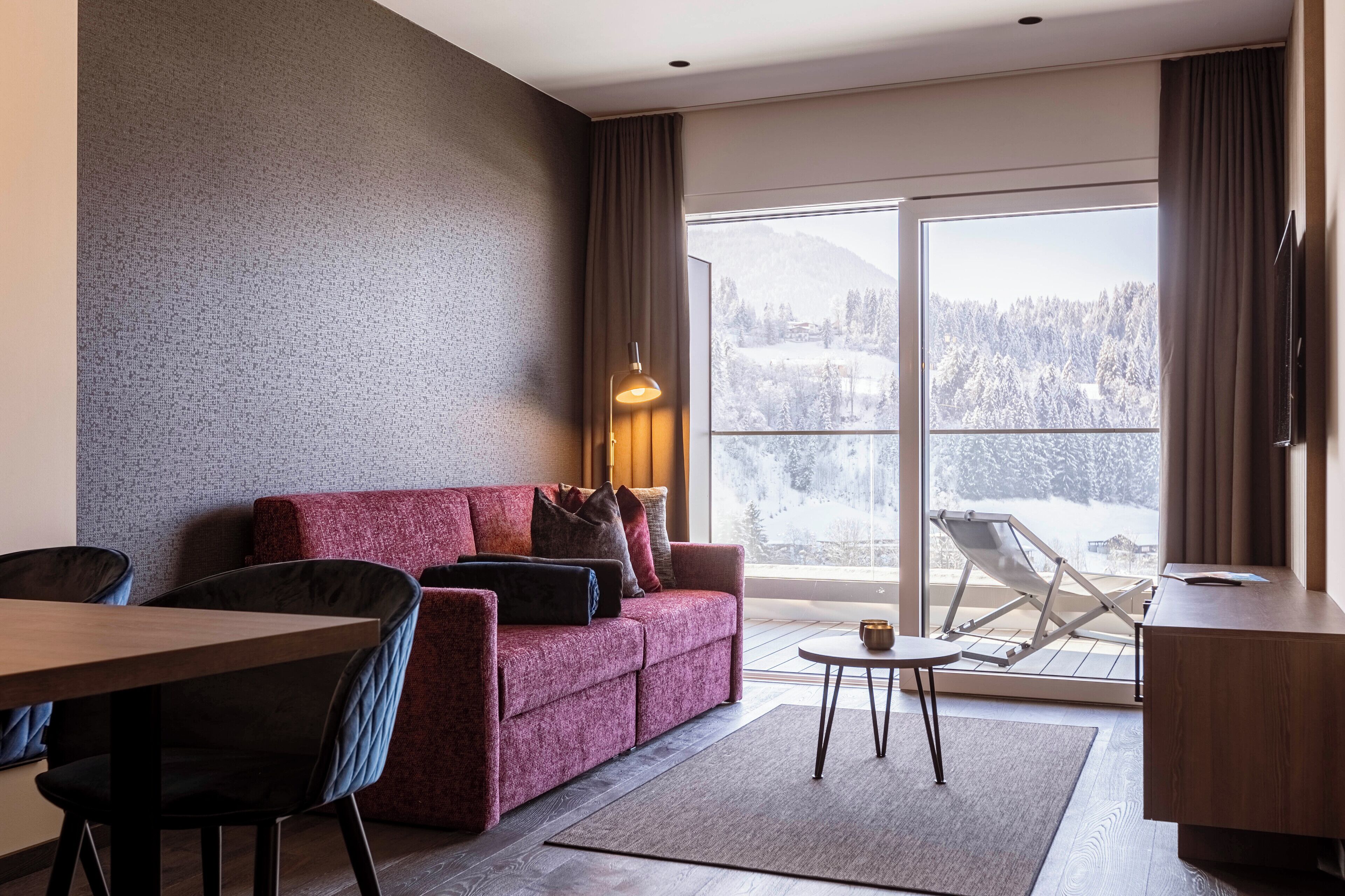 A modern living room with a red sofa, a wooden table, and a balcony with a view of snow-covered mountains.