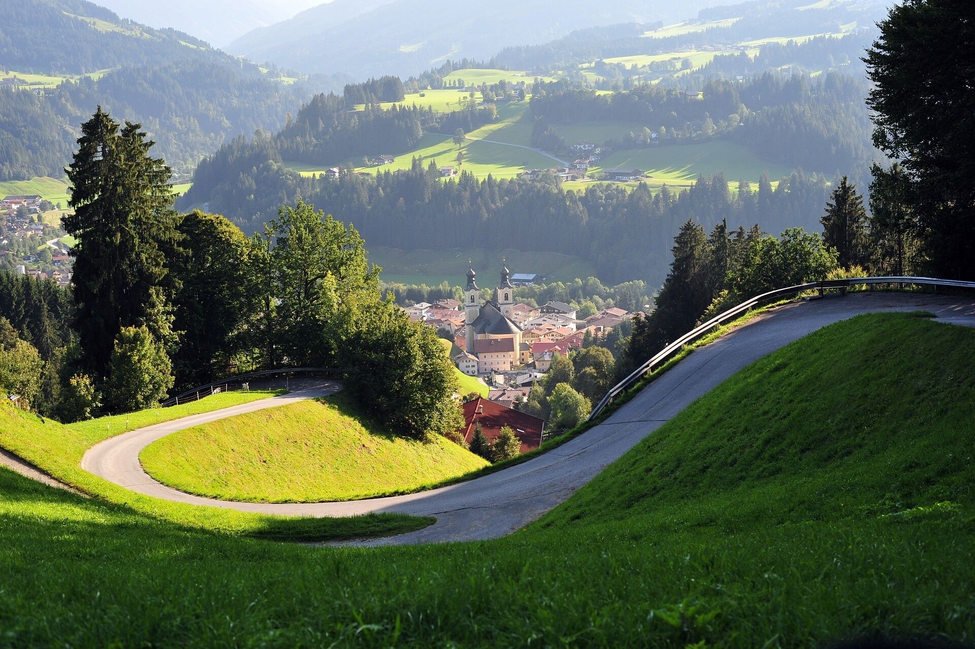 Une route sinueuse traverse un paysage verdoyant, avec une ville et une église visibles au loin.