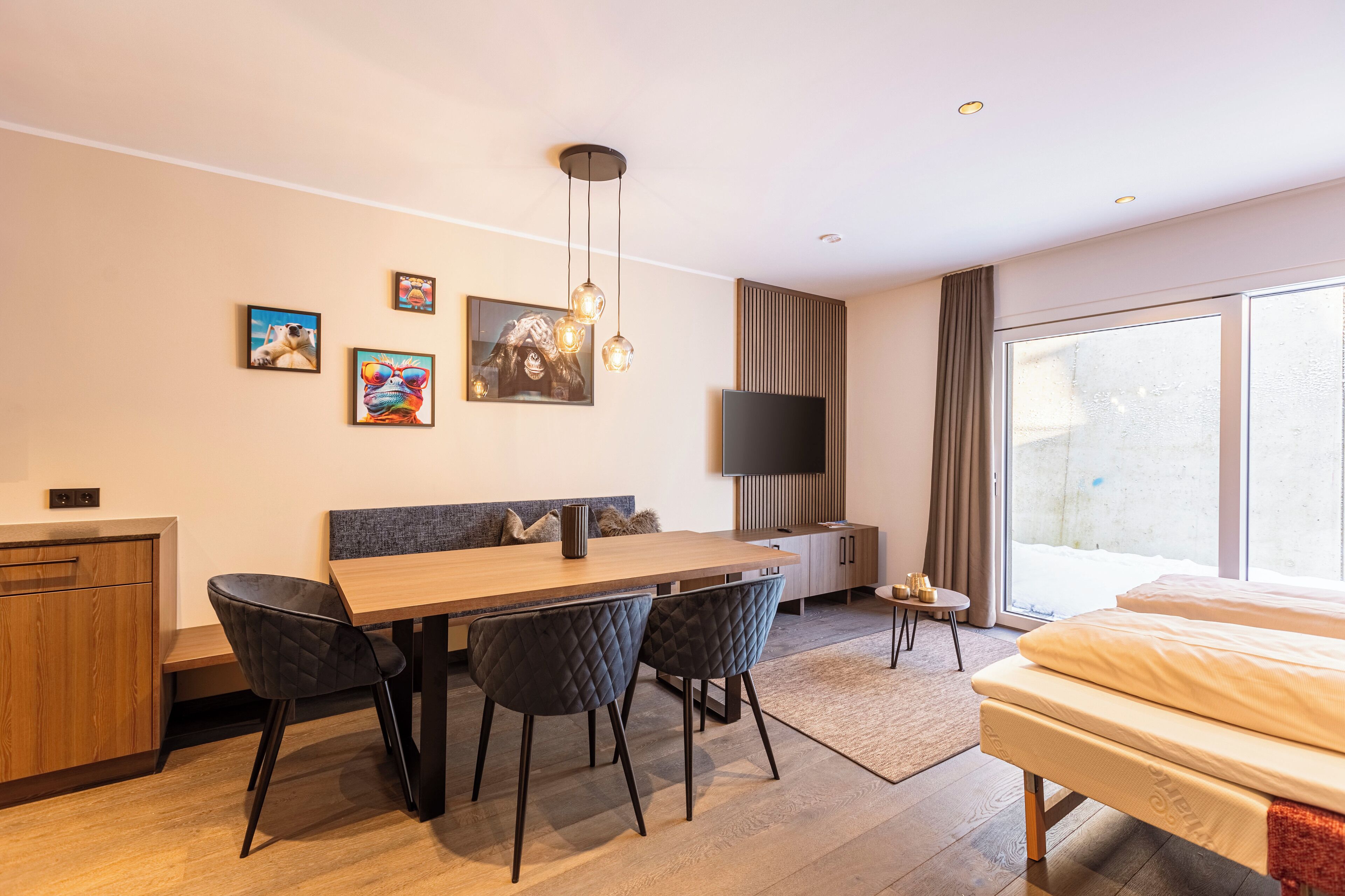 Una habitación de hotel moderna con una mesa de comedor de madera, sillas negras, un televisor de pantalla plana, un ventanal grande y una cama.