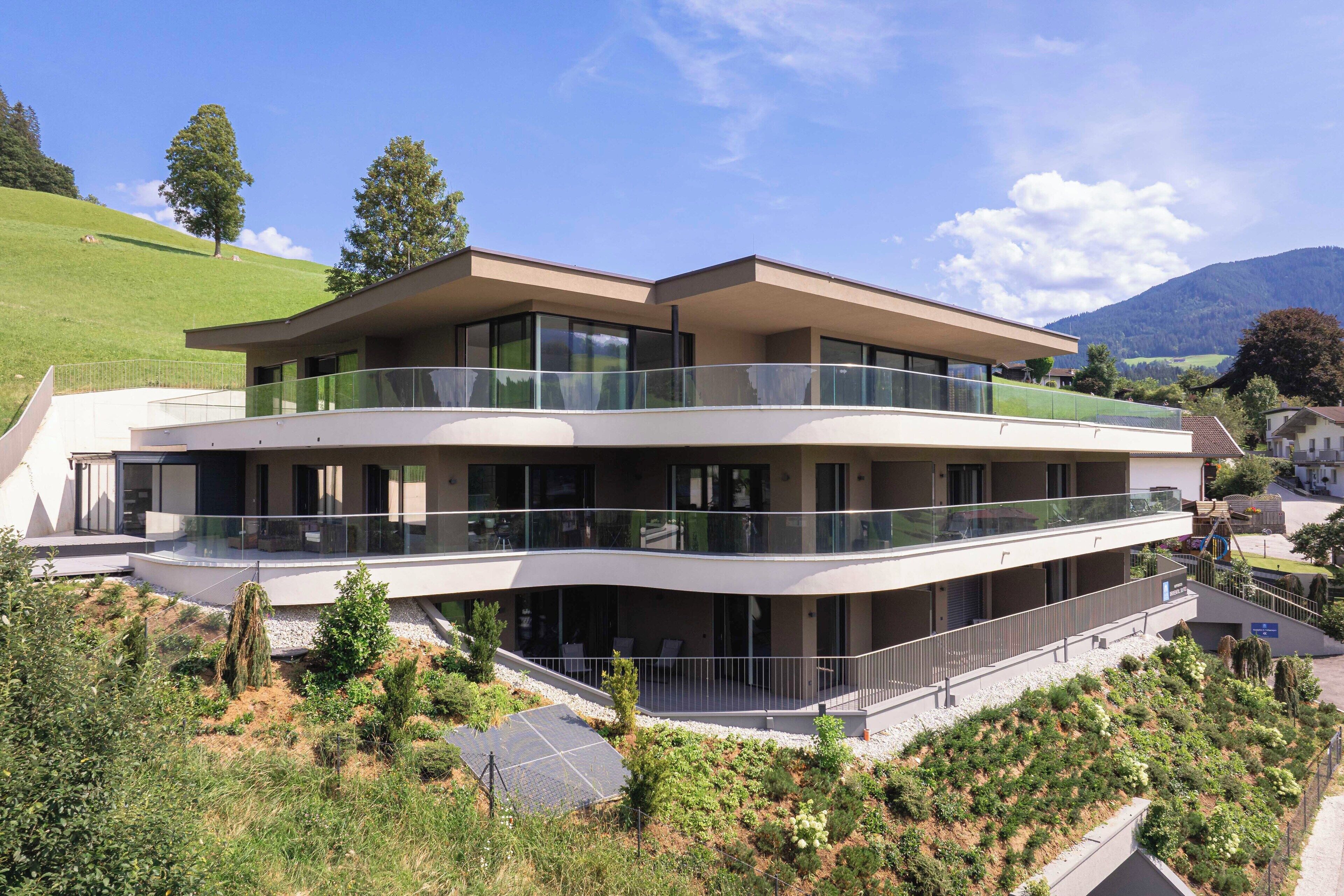 A modern multi-story building with a glass balcony, surrounded by greenery and a hilly landscape.