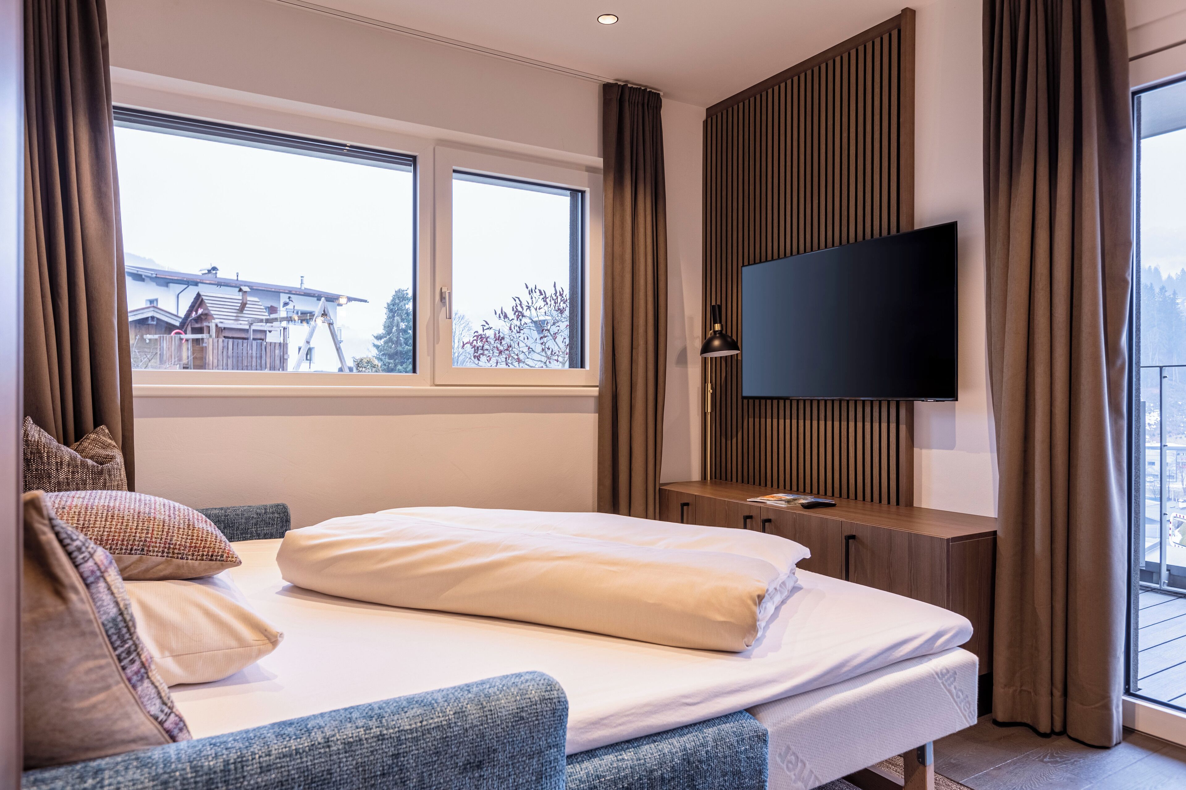 A modern hotel room with a large bed, a TV mounted on the wall, and a balcony with a view of the outside.