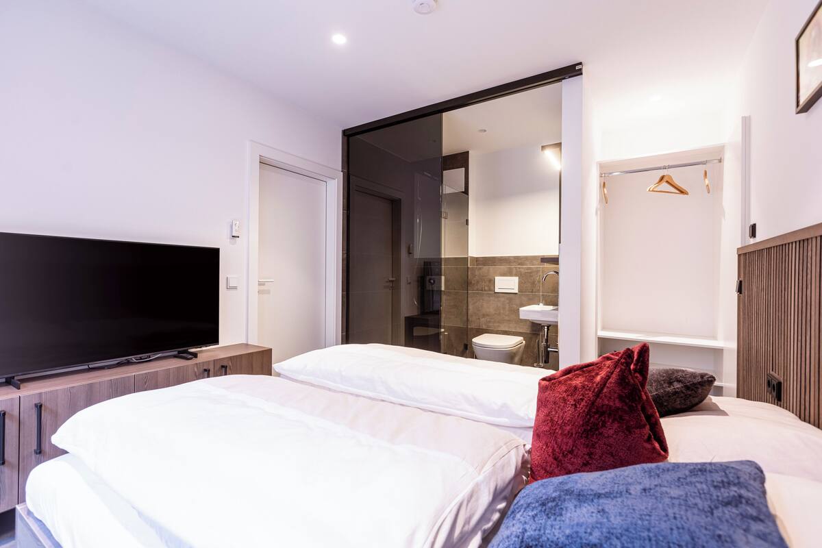 Moderni hotellihuone, jossa on suuri litteänäyttöinen televisio, kaksi sänkyä, liukuovi kylpyhuoneeseen ja puinen pääty.