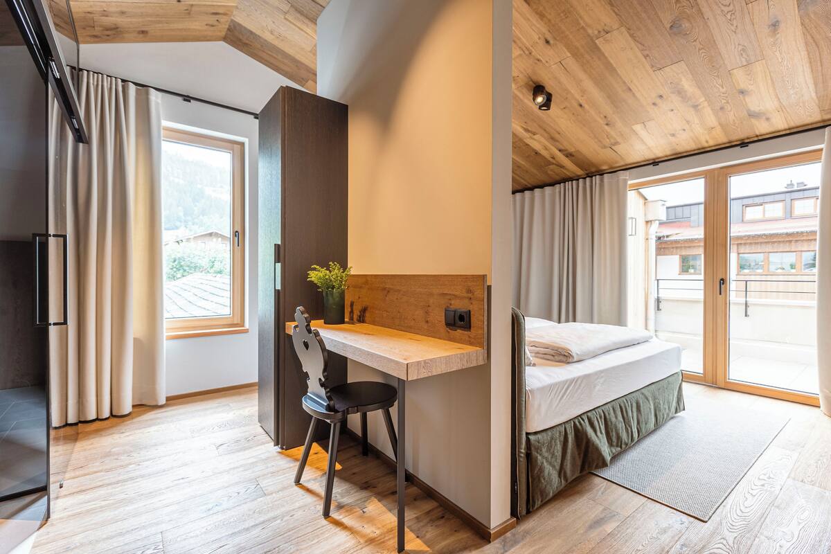 Ein Schlafzimmer mit Holzboden, einem Schreibtisch, einem Stuhl, einem Bett und Blick auf Gebäude durch große Fenster.