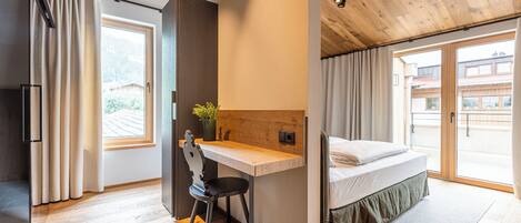 Une chambre avec un plafond en bois, un bureau, une chaise, un lit et une vue sur les bâtiments à travers de grandes fenêtres.