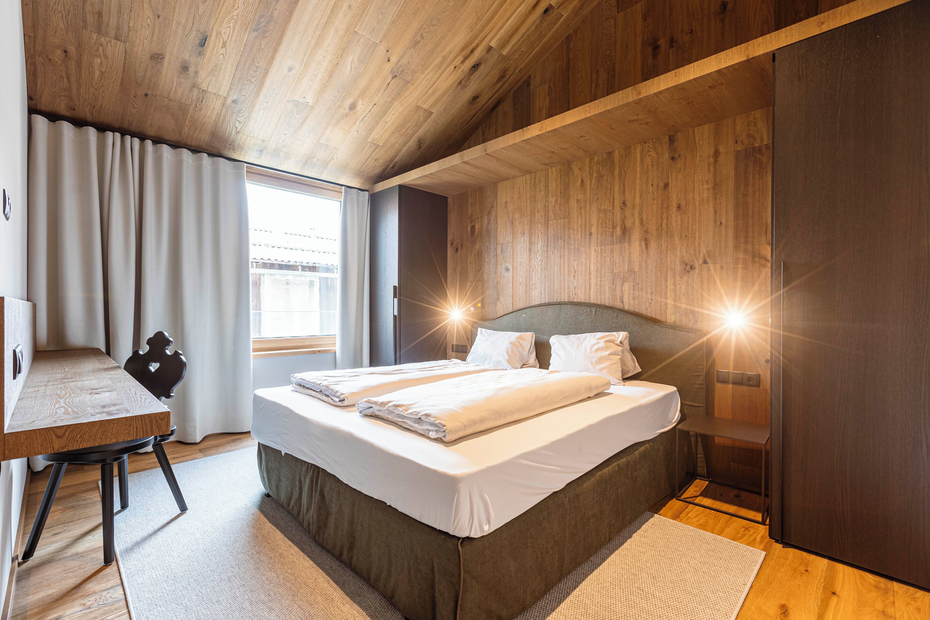 Quarto moderno com teto de madeira, uma cama grande com roupa de cama branca, uma escrivaninha e uma janela com cortinas.