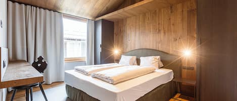 Quarto moderno com teto de madeira, uma cama grande com roupa de cama branca, uma escrivaninha e uma janela com cortinas.
