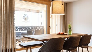Un coin repas avec une table en bois, quatre chaises, un luminaire suspendu et une vue sur l’extérieur grâce à de grandes fenêtres.