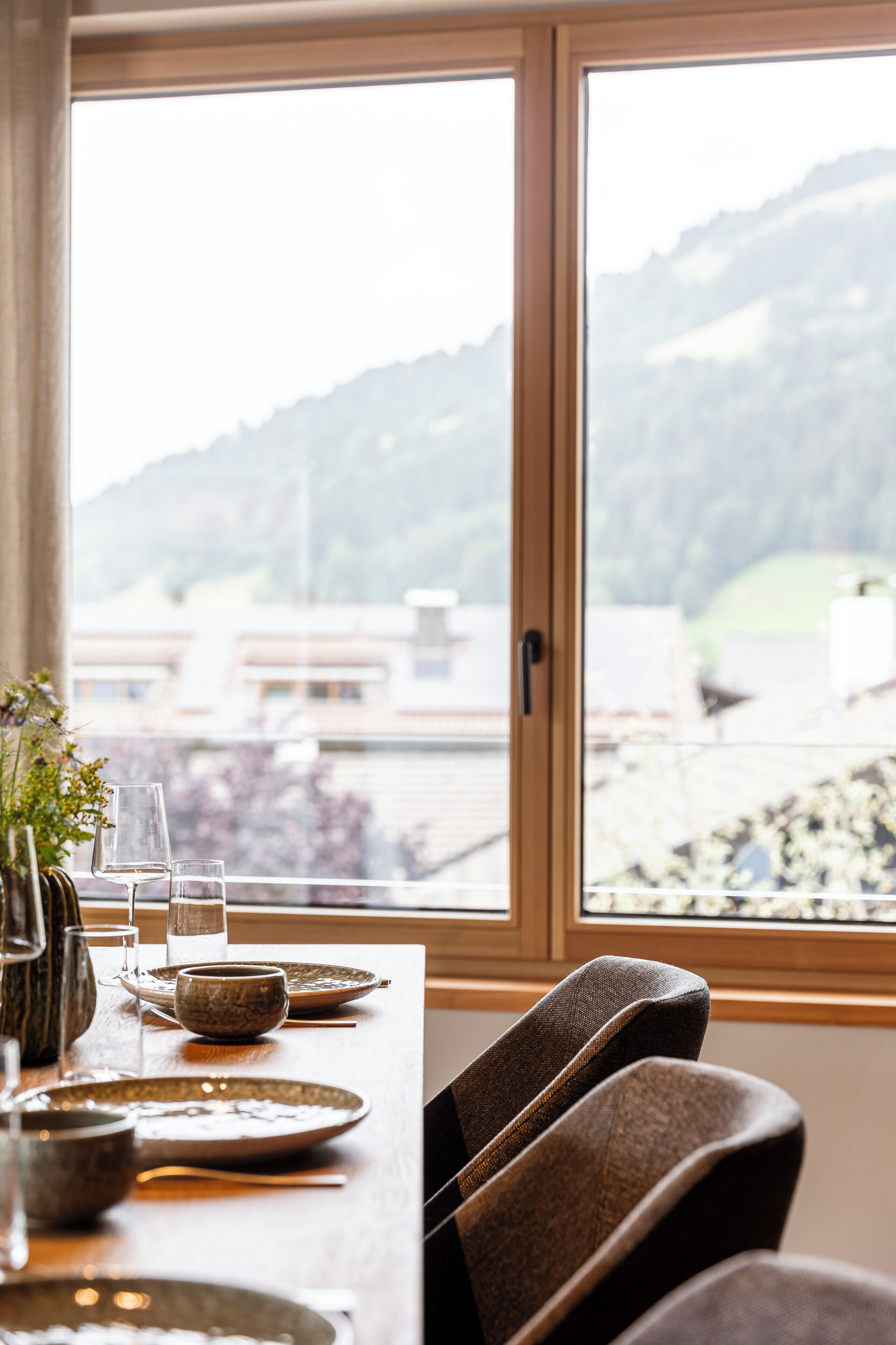 A dining table with plates, bowls, and glasses, set against a window with a view of buildings and hills.