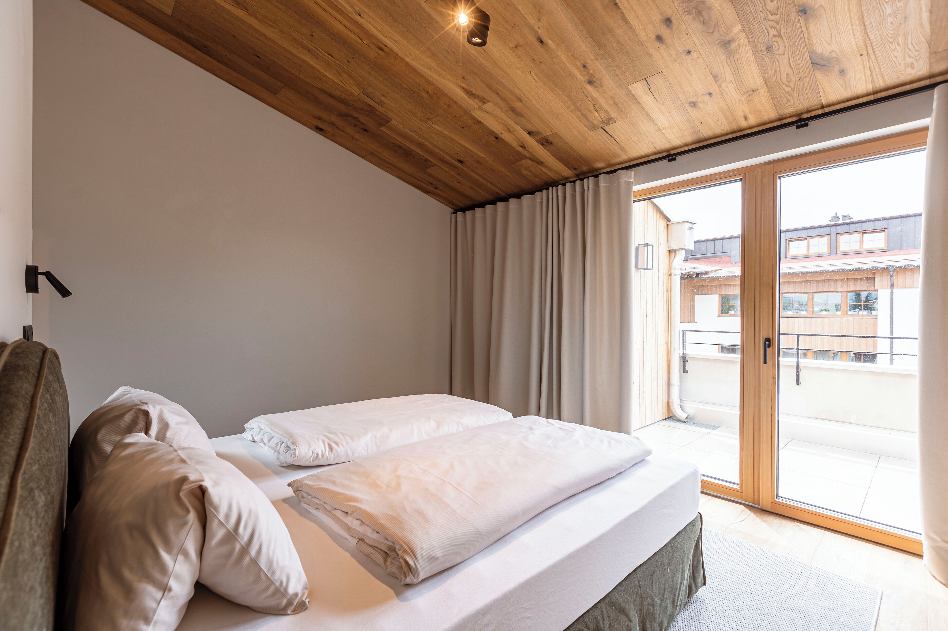 Une chambre d’hôtel avec un plafond en bois, un lit recouvert de draps blancs, une vue sur l’extérieur à travers une porte-fenêtre coulissante et une fenêtre avec des rideaux.