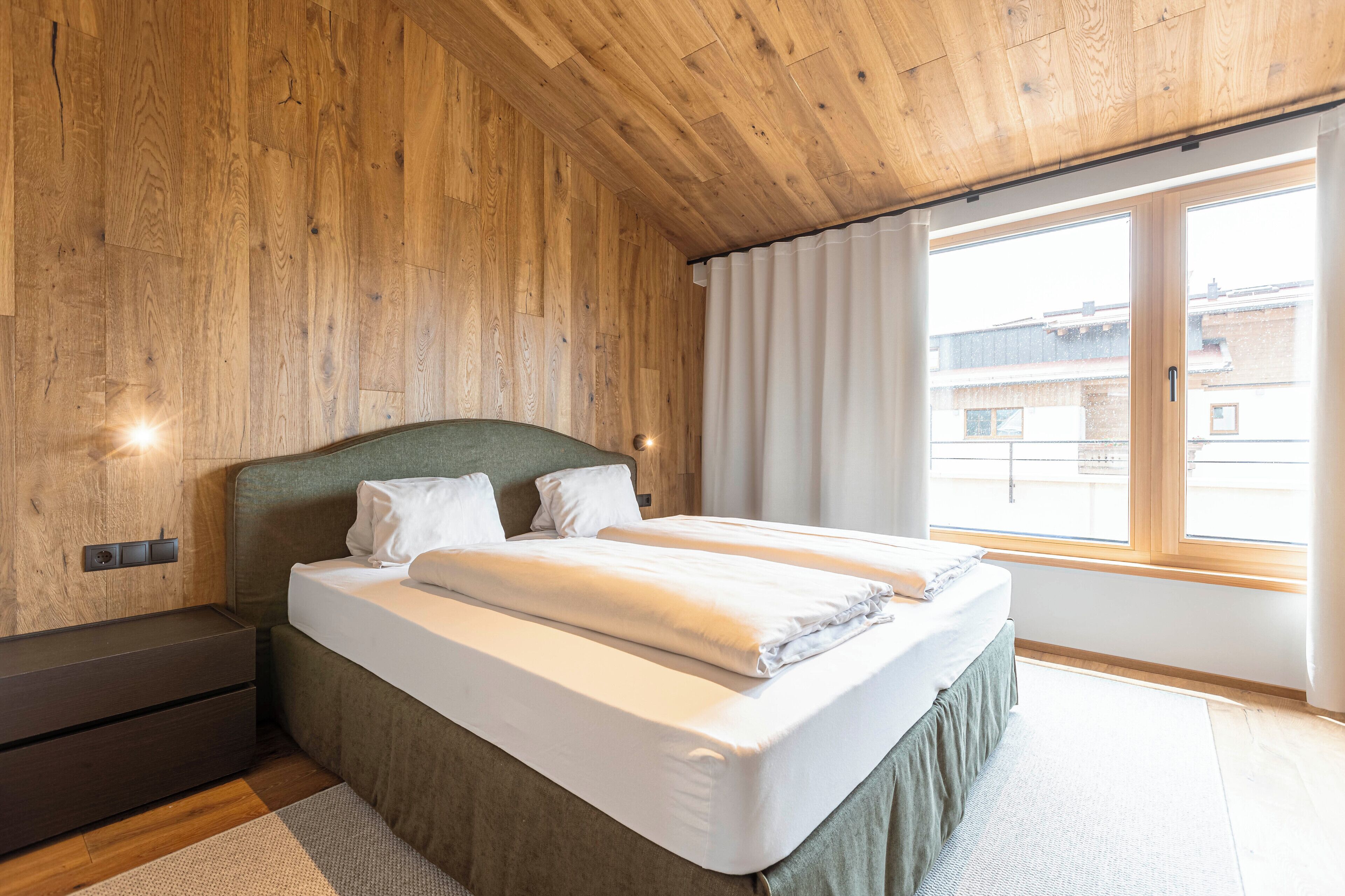 Une chambre moderne avec un plafond en bois, une grande fenêtre et un lit recouvert de draps blancs.