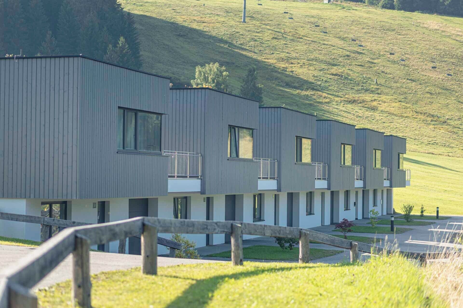 A modern building with a flat roof, multiple balconies, and a grassy hill in the background.
