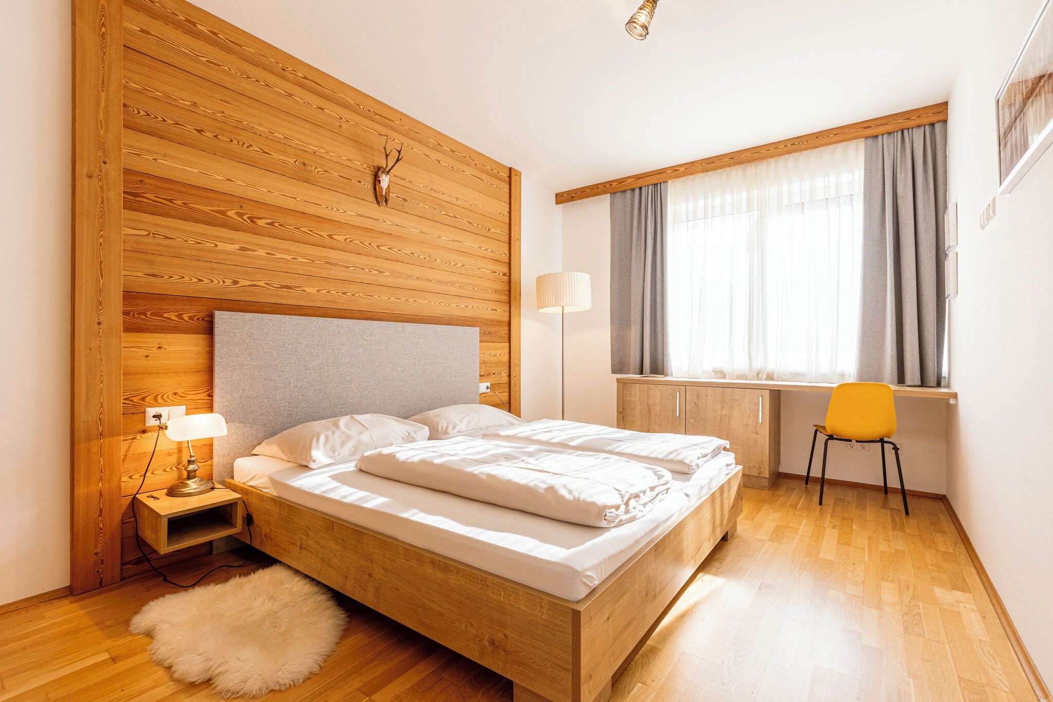 Ein Schlafzimmer mit Holzkopfende, einem Bett mit weißer Bettwäsche, einem Nachttisch mit Lampe, einem Sessel und einem Fenster mit Vorhängen.