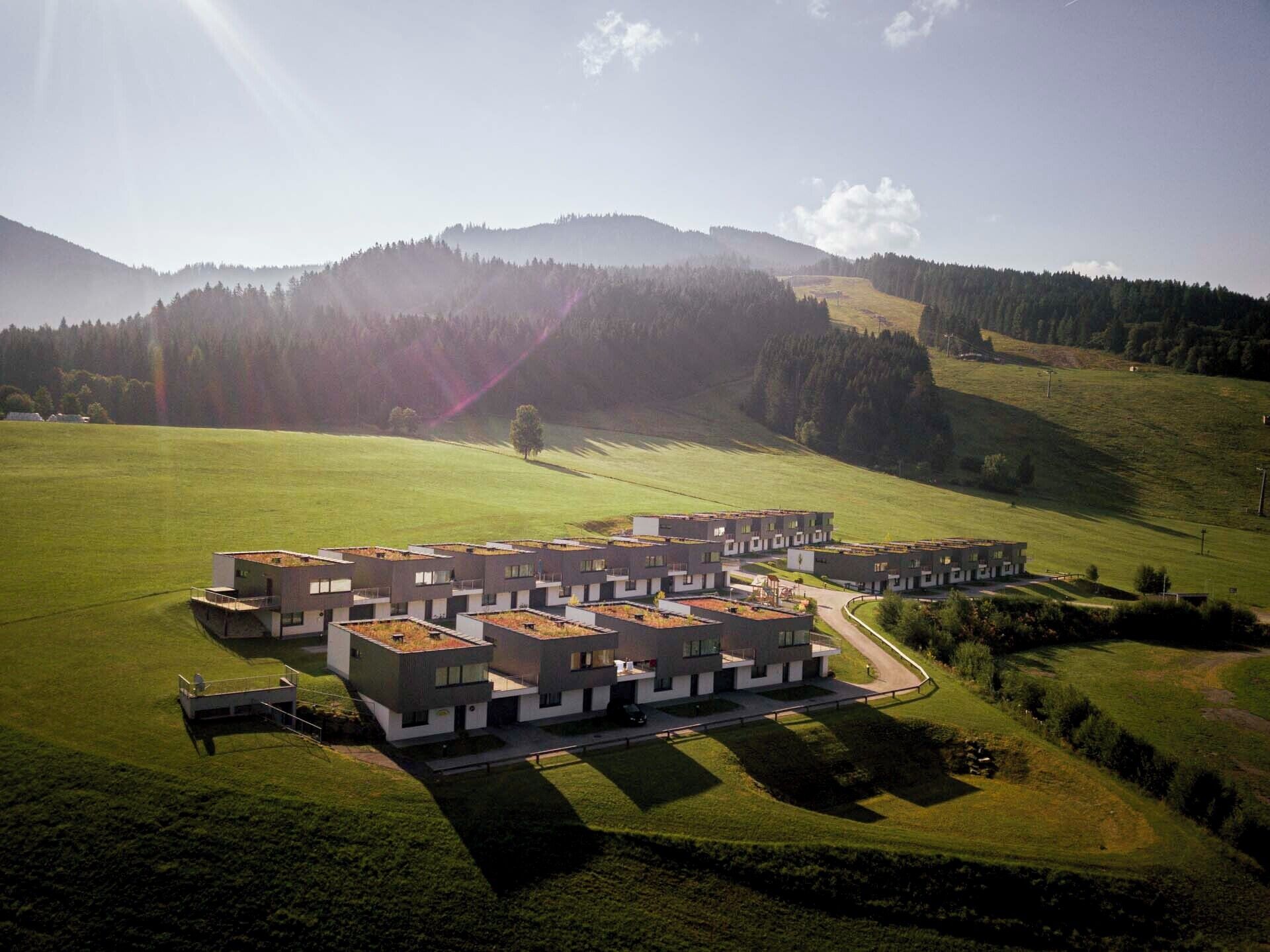 A modern residential complex with multiple buildings, a central courtyard, and surrounding green fields and hills.