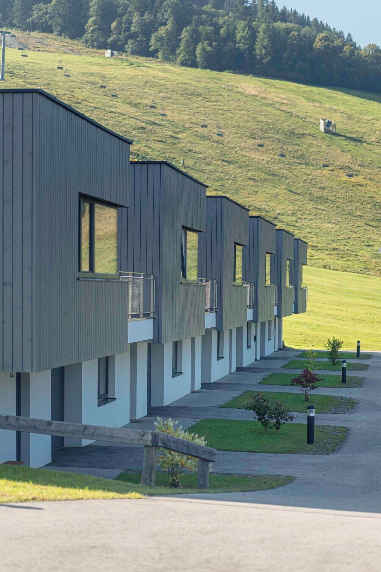 Une rangée de bâtiments modernes à un seul étage, dotés de grandes fenêtres et de balcons, s’étend sur une zone verdoyante, avec une colline en arrière-plan.