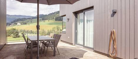An outdoor patio with a table and chairs, a lounge chair, and a view of a green landscape.