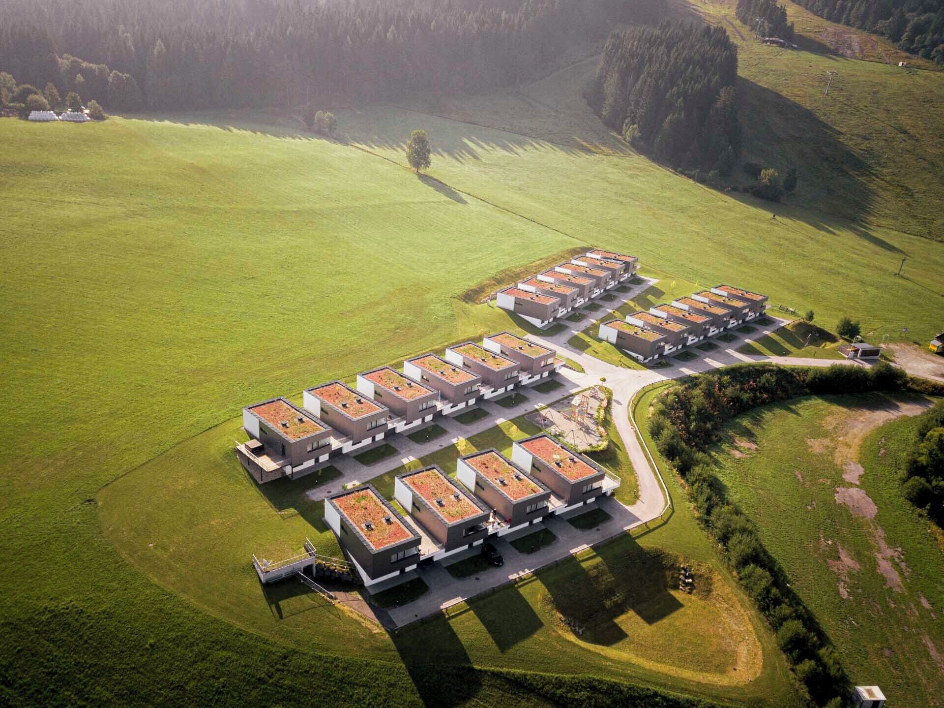 An aerial view of a modern residential complex with multiple buildings arranged in a grid pattern, surrounded by green fields and trees.