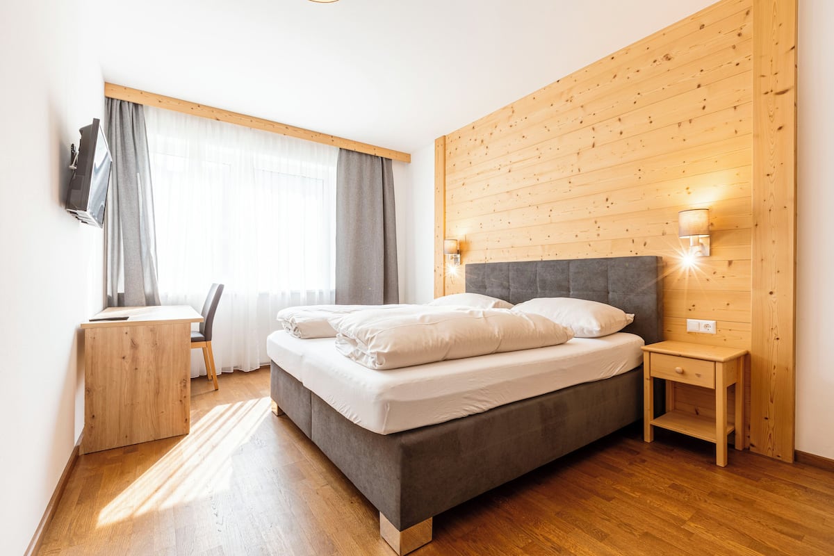A modern bedroom with a large bed, a wooden headboard, a desk with a chair, and a window with curtains.