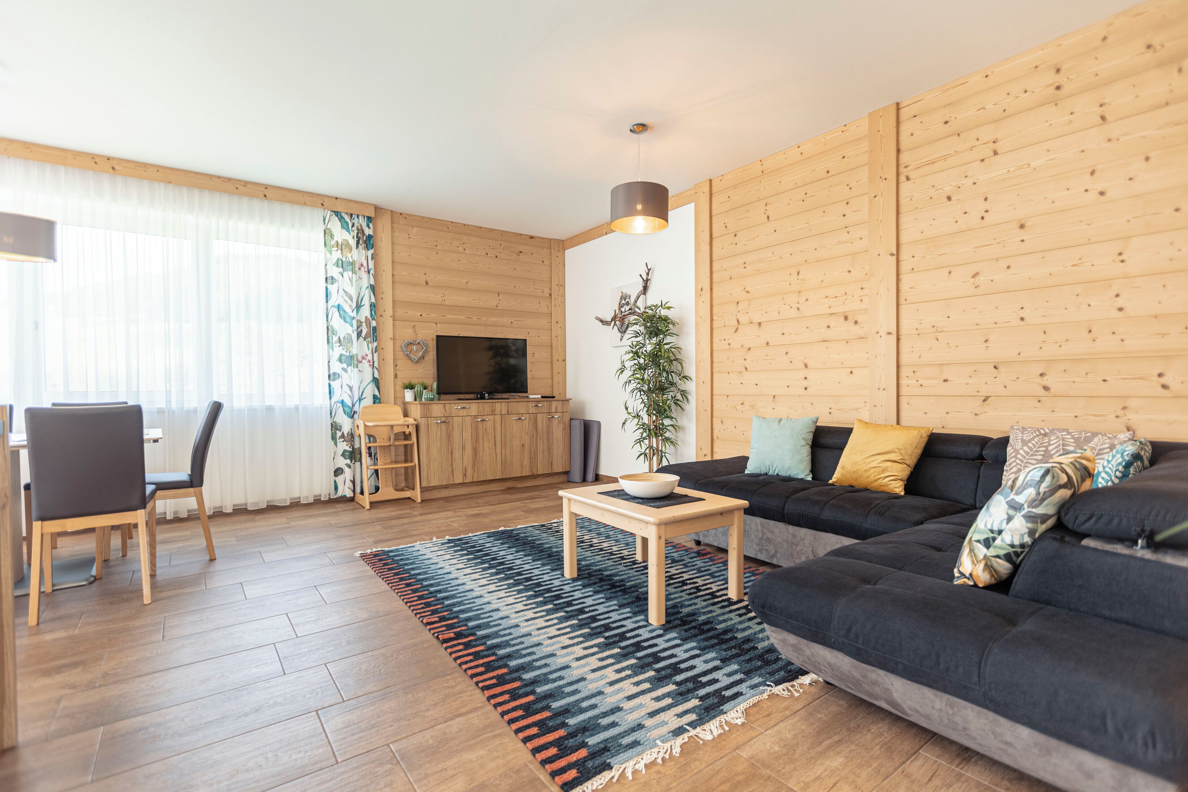 A modern living room with a dark sofa, wooden flooring, a television, and a dining area with chairs and a table.