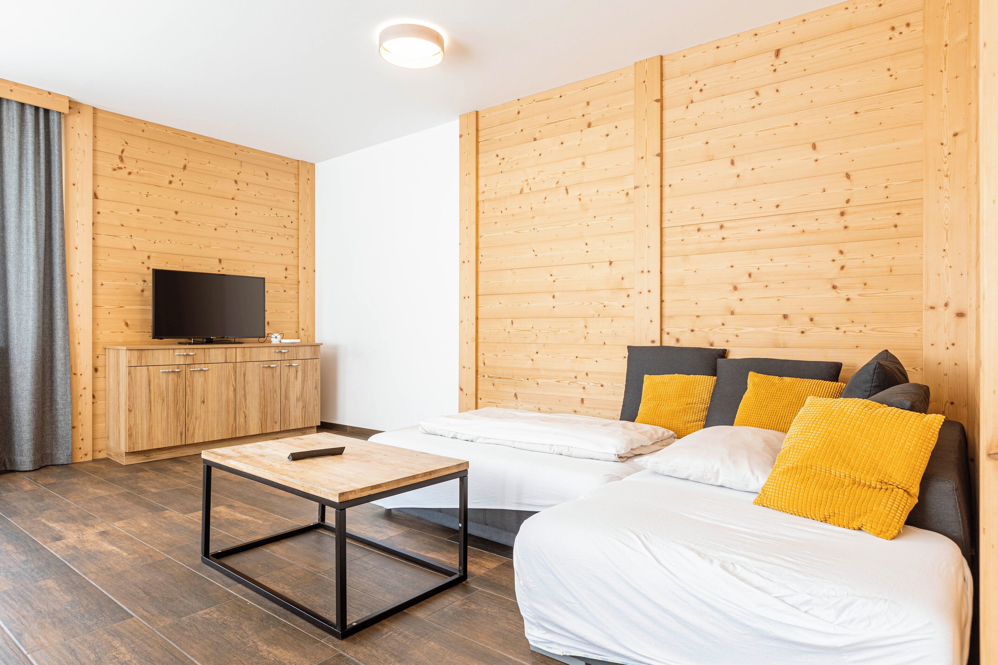 Una camera moderna con pareti rivestite in legno, un televisore a schermo piatto su un mobile in legno, un letto con biancheria da letto bianca e cuscini gialli, e un tavolino con un telecomando.