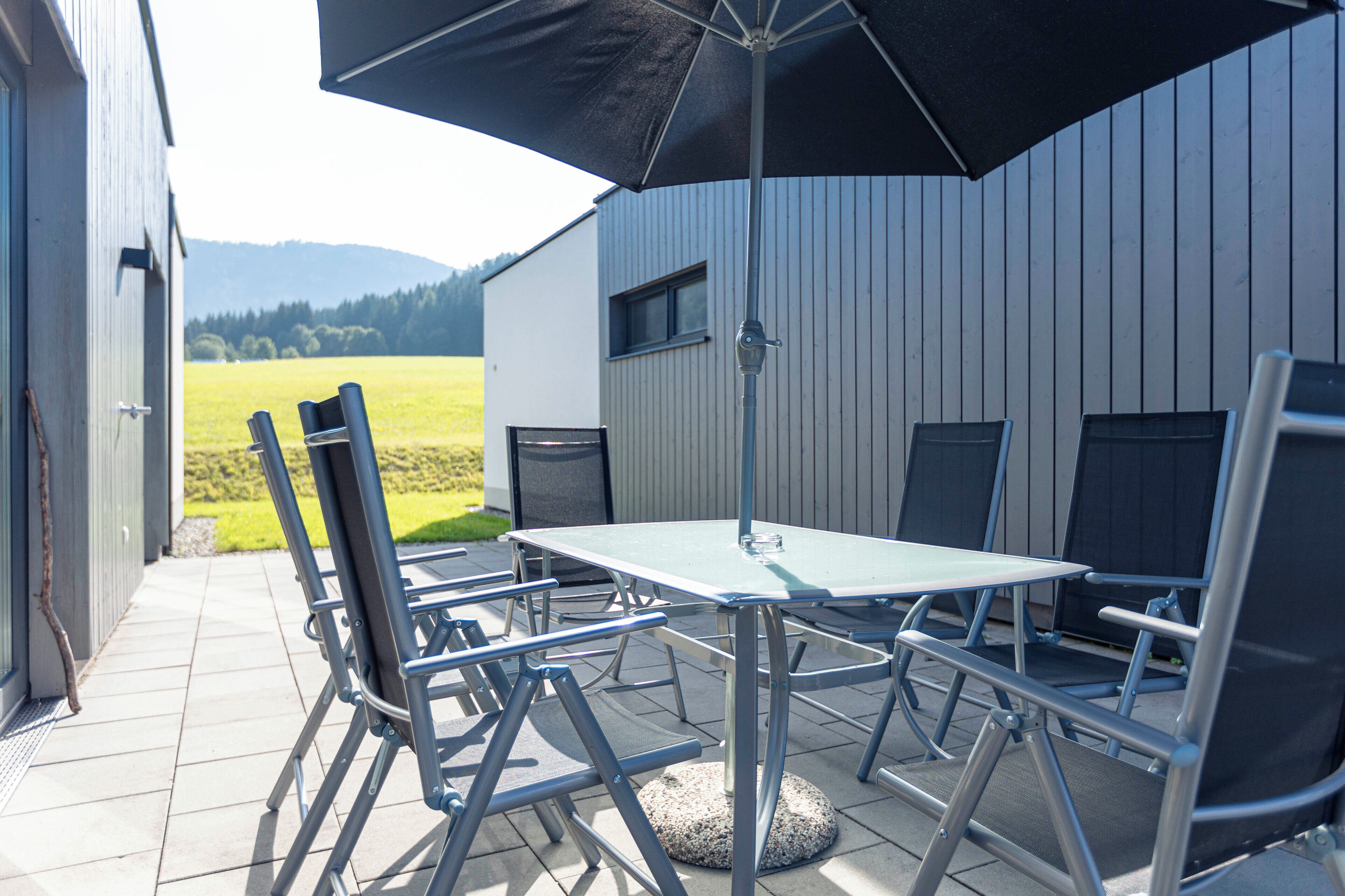 Eine Terrasse im Freien mit Tisch und Stühlen, einem großen Sonnenschirm und Blick auf ein Feld und Berge.