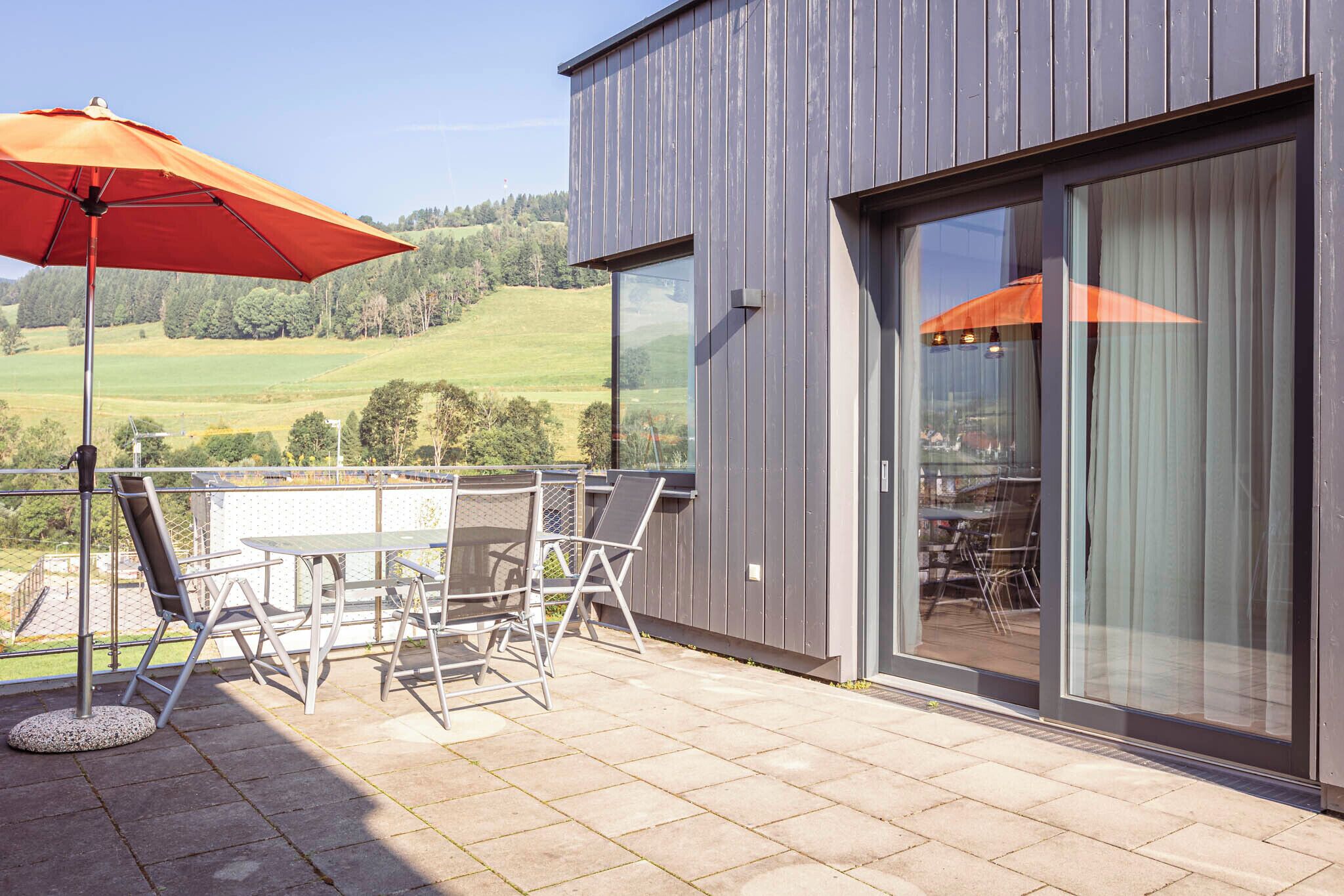 Ein modernes Gebäude mit einem Balkon, der einen Tisch, Stühle und einen orangefarbenen Sonnenschirm bietet, mit schöner Aussicht auf eine grüne Landschaft.
