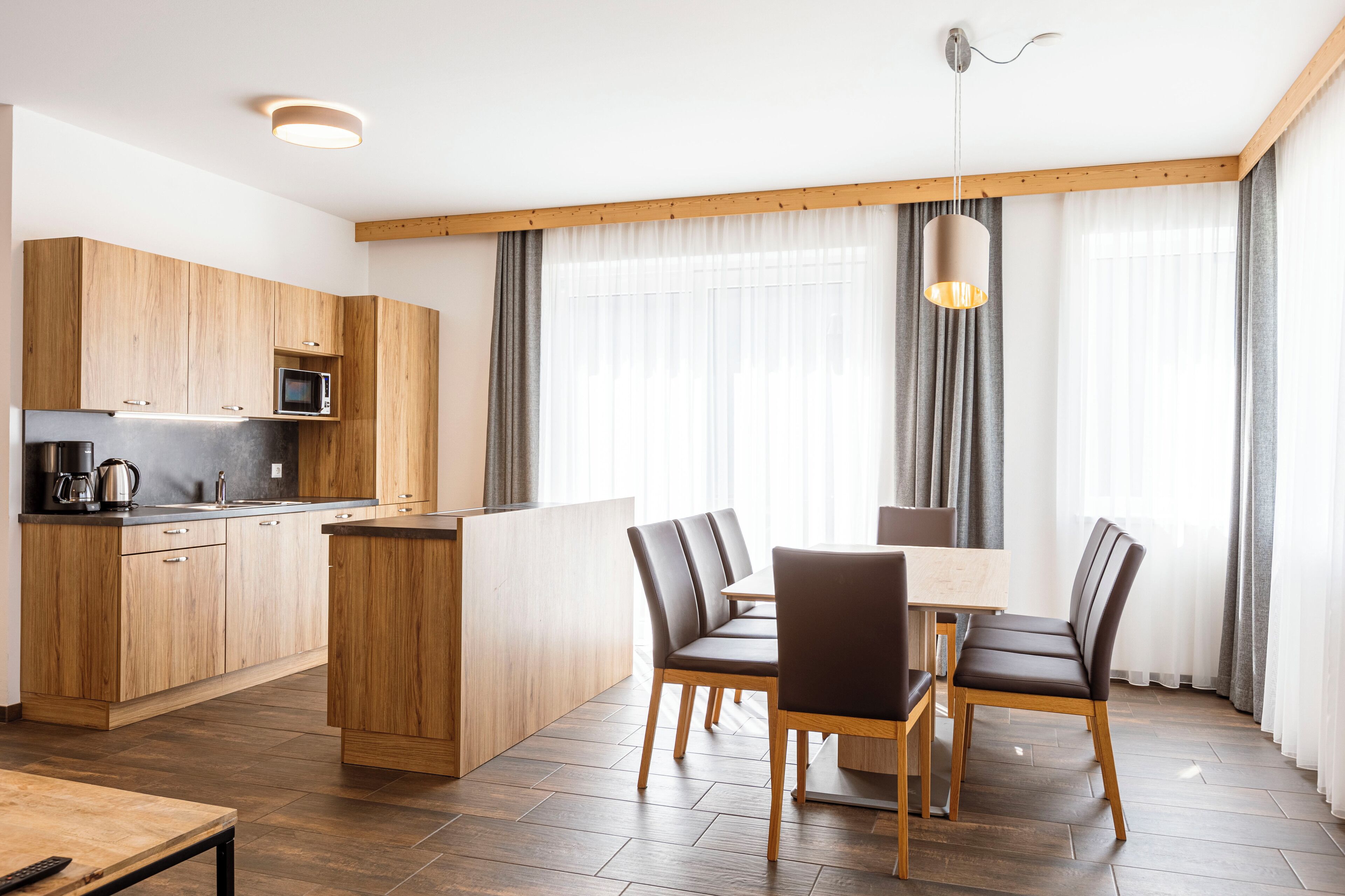 Eine moderne Küche mit Holzausstattung, einer Mikrowelle und einer Kaffeemaschine. Es gibt eine Essecke mit Tisch und Stühlen sowie ein großes Fenster mit Vorhängen.