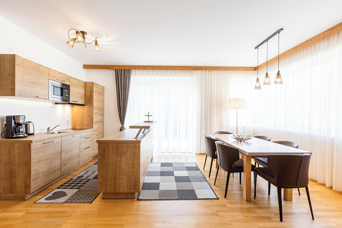 A modern kitchen with wooden cabinets, a built-in microwave, and a coffee maker. There is a dining area with a table and chairs, and a large window with sheer curtains.