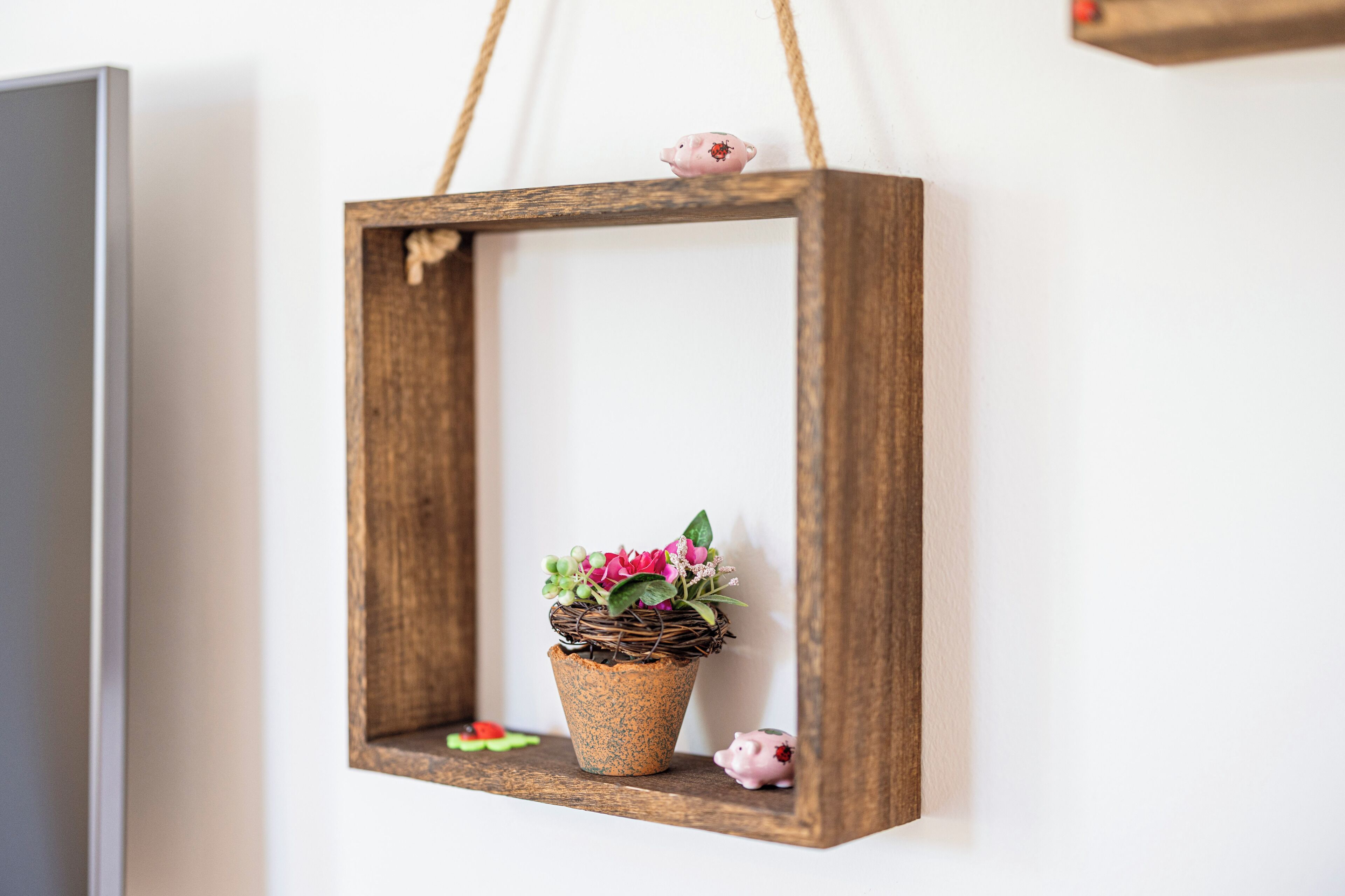 Ein rustikaler Holzrahmen mit einem gewebten Korb, der rosa Blüten enthält, hängt an einer weißen Wand.