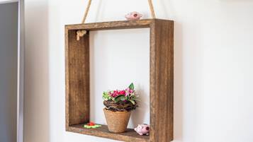 Ein rustikaler Holzrahmen mit einem gewebten Korb, der rosa Blüten enthält, hängt an einer weißen Wand.