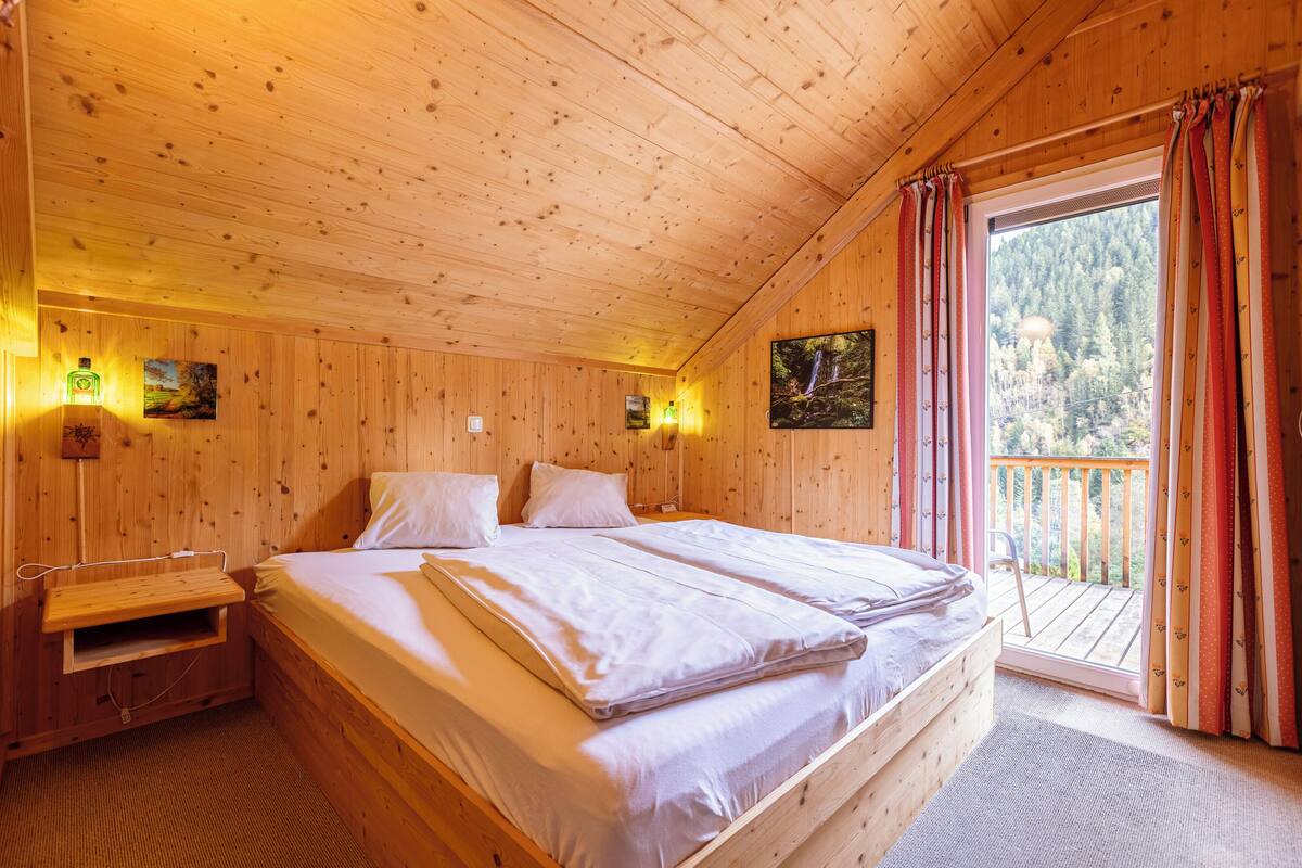 Habitación de cabaña de madera con cama, un pequeño banco y un balcón con vistas a árboles.