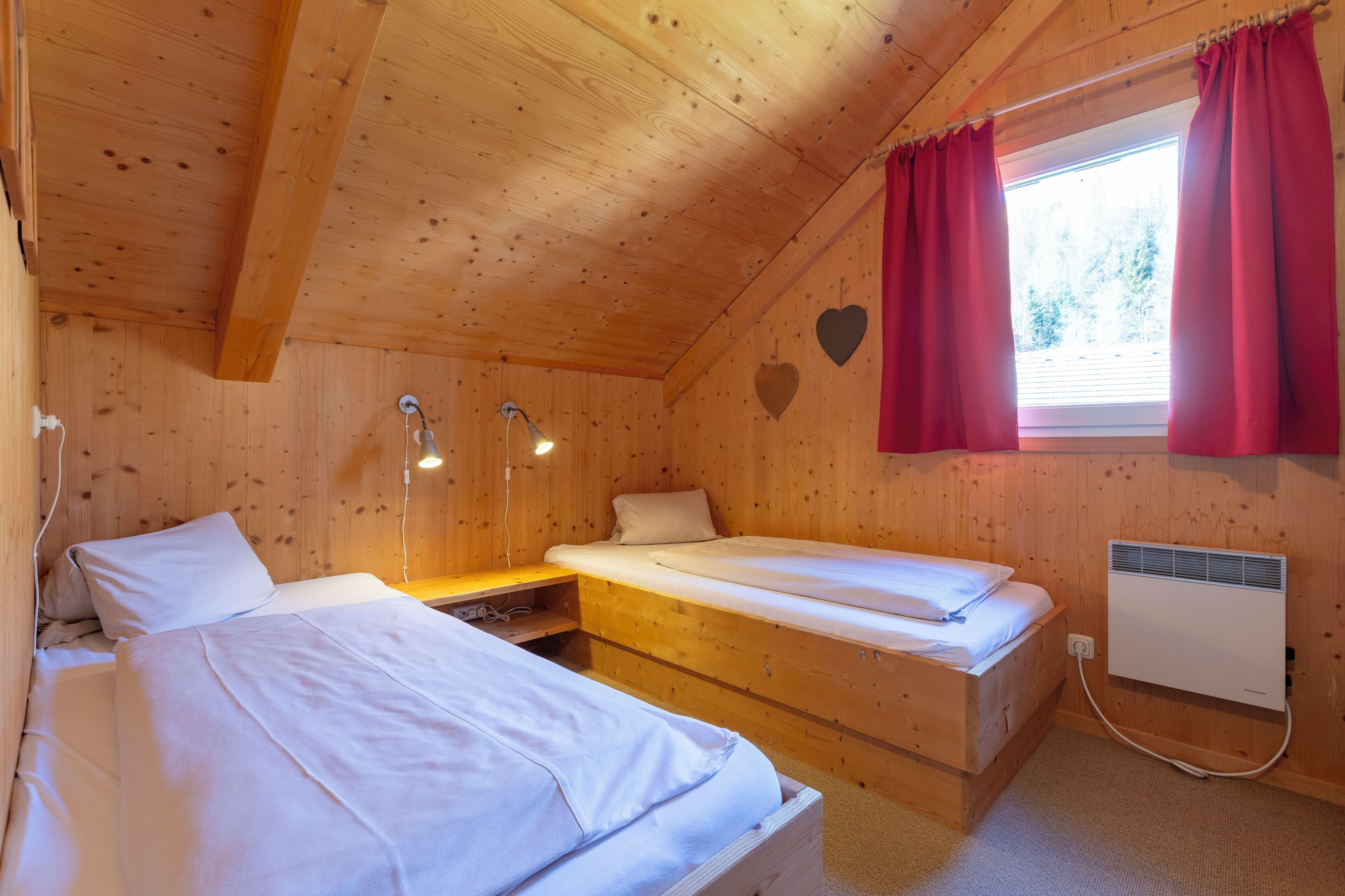 Ein Zimmer in einer Holzhütte mit zwei Betten, einem Fenster mit roten Vorhängen, einem Wandheizgerät und zwei Nachttischlampen.