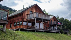 Une cabane en bois dotée d’un balcon et d’une terrasse au deuxième étage, entourée d’une colline verdoyante.