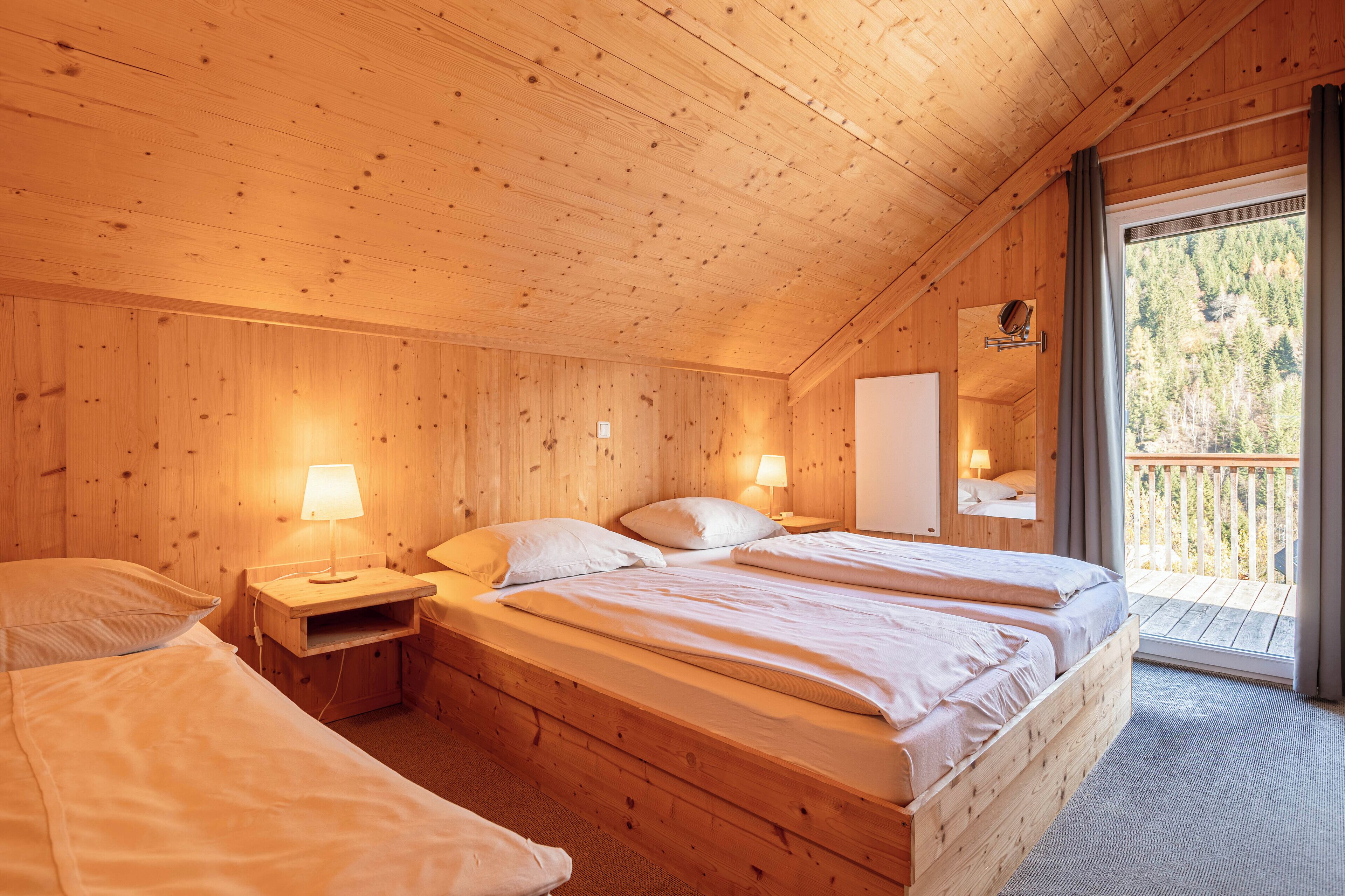 Habitación de cabaña de madera con dos camas, una mesita de noche, un espejo y una ventana con vistas a árboles.