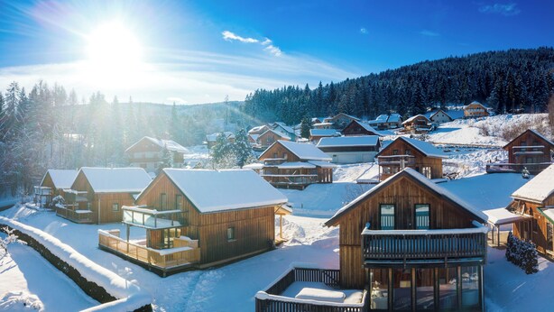 Une station de ski enneigée, avec des chalets en bois, un ciel ensoleillé et une forêt en arrière-plan.