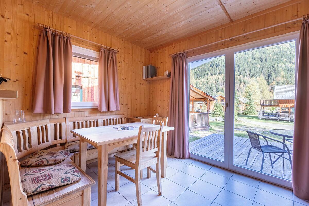 Interior de una cabaña de madera con una mesa de comedor, sillas, un banco con cojines y un ventanal corredero que da a una terraza exterior.