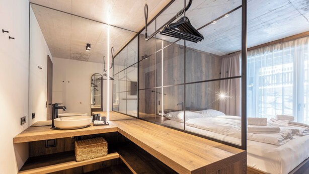 Een moderne hotelkamer met een houten wastafel, een grote spiegel, een bed met witte lakens en een stadszicht door grote ramen.