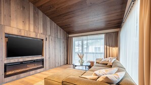 Een moderne woonkamer met een grote flatscreen televisie, houten lambrisering, een bank met kussens, een open haard en een groot raam met gordijnen.