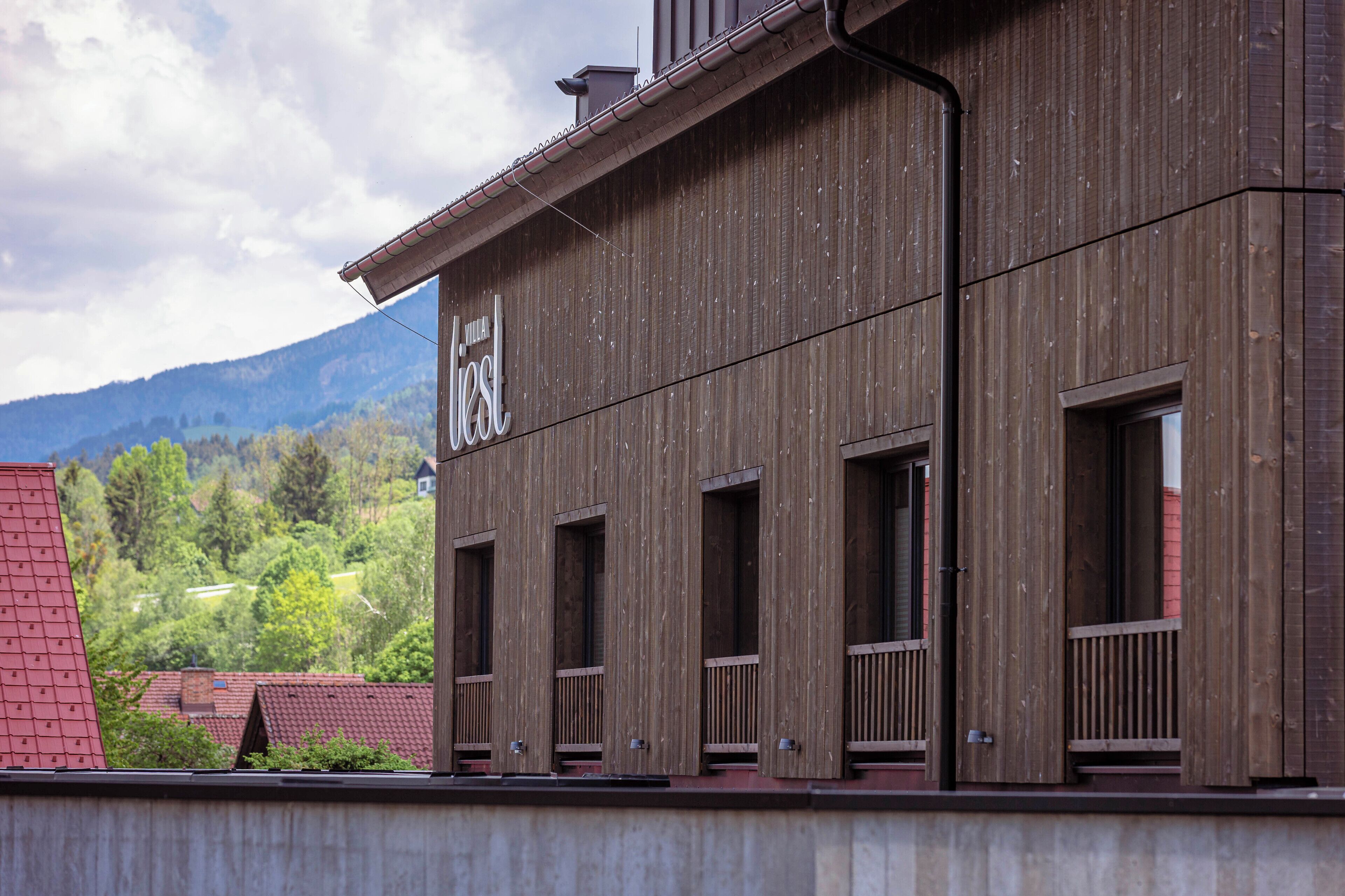 A modern building with a wooden facade, multiple windows, and a view of a green landscape with mountains in the background.
