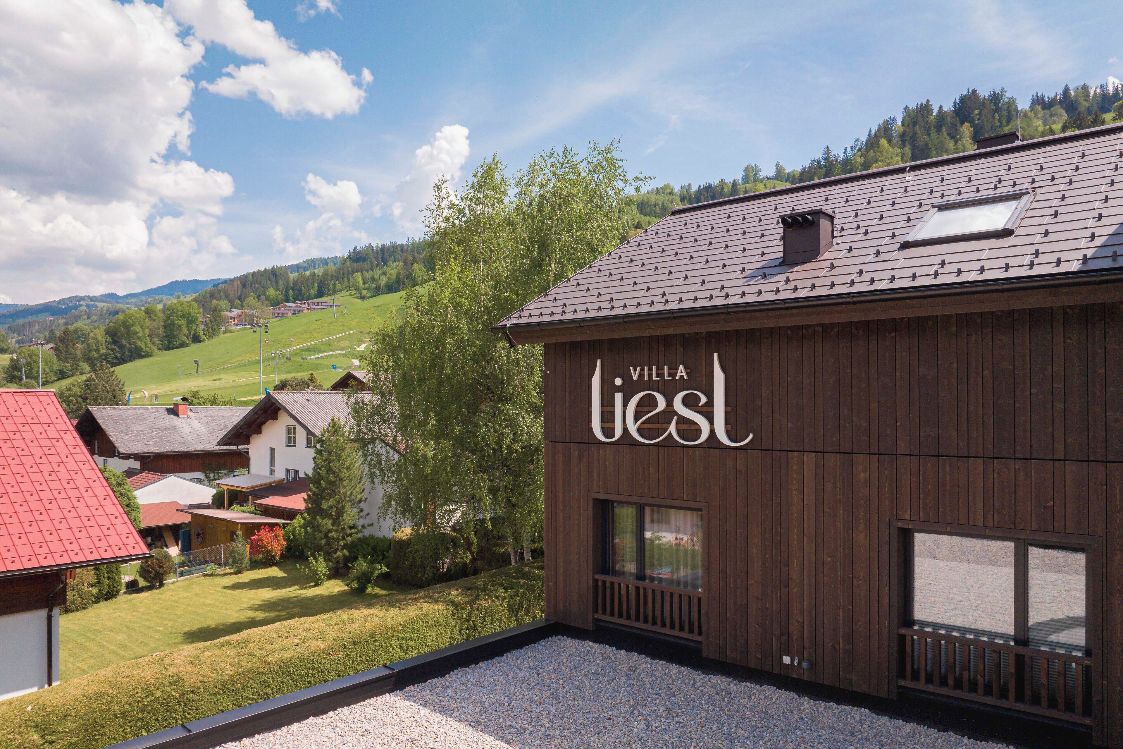 A villa with a wooden exterior, a gravel patio, and a view of a green valley and houses in the background.