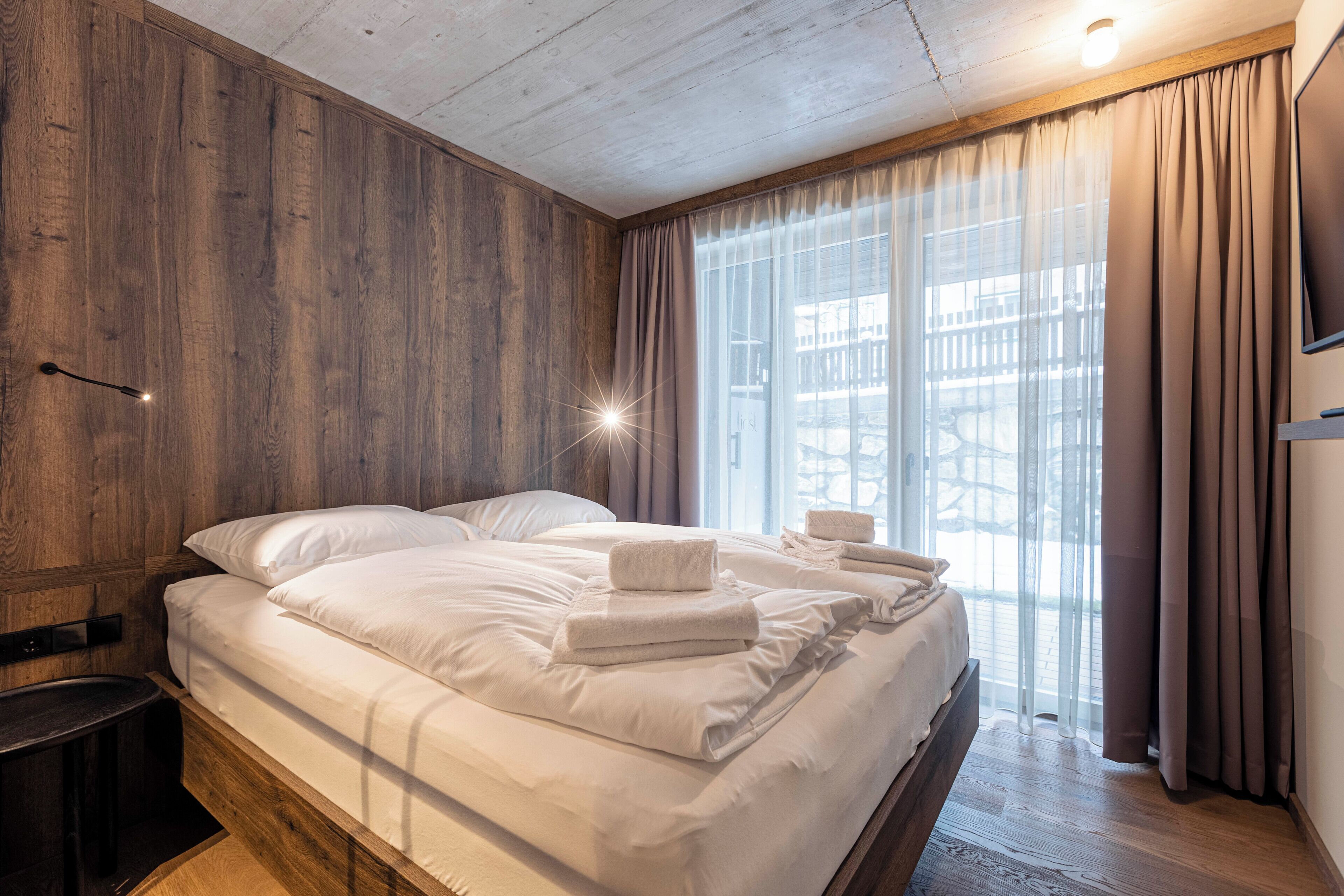 A double bed with white linens and towels, wooden paneling on the walls, a nightstand, and a large window with curtains.