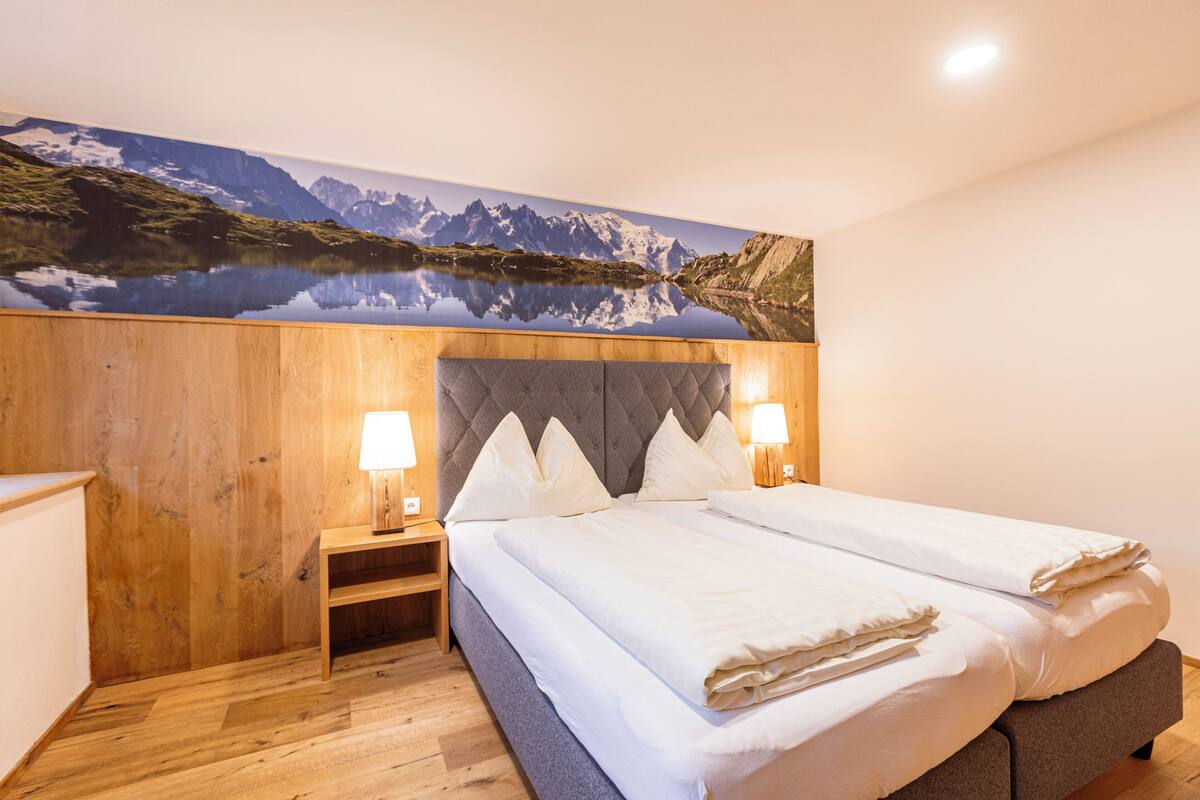 Quarto com cabeceira de madeira, duas mesas de cabeceira com abajures, uma cama grande com roupa de cama branca e uma pintura de paisagem montanhosa na parede.