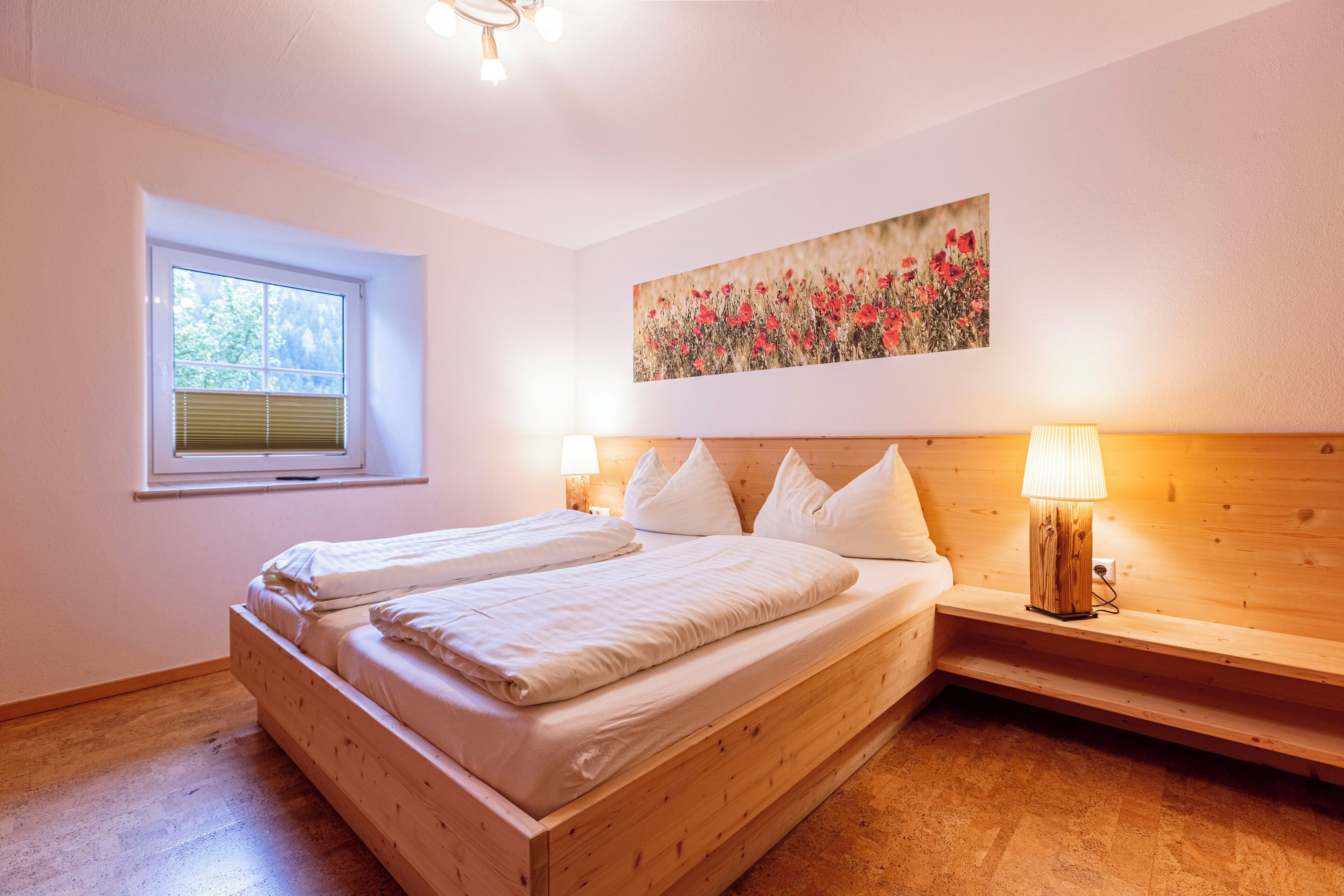 Un dormitorio con cama de madera, ventana con persianas, un cuadro en la pared y una lámpara de noche.