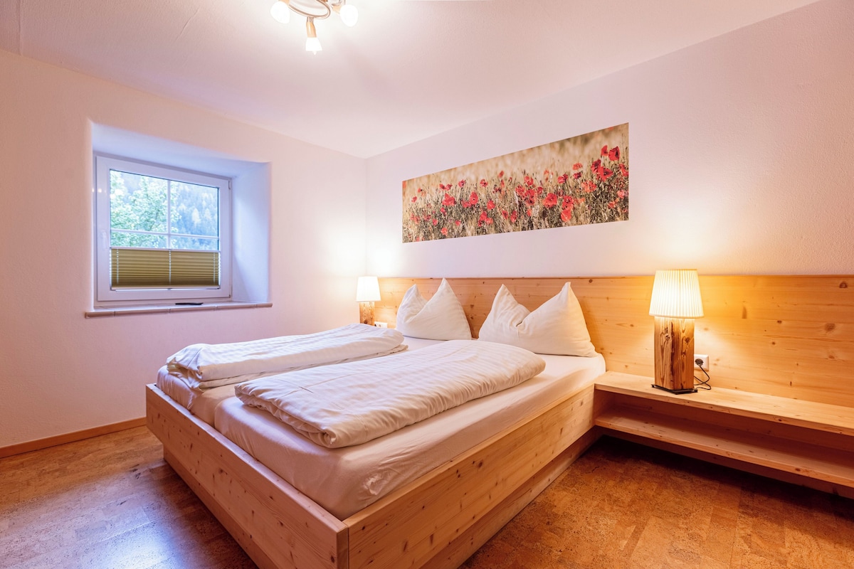 Un dormitorio con cama de madera, ventana con persianas, un cuadro en la pared y una lámpara de noche.