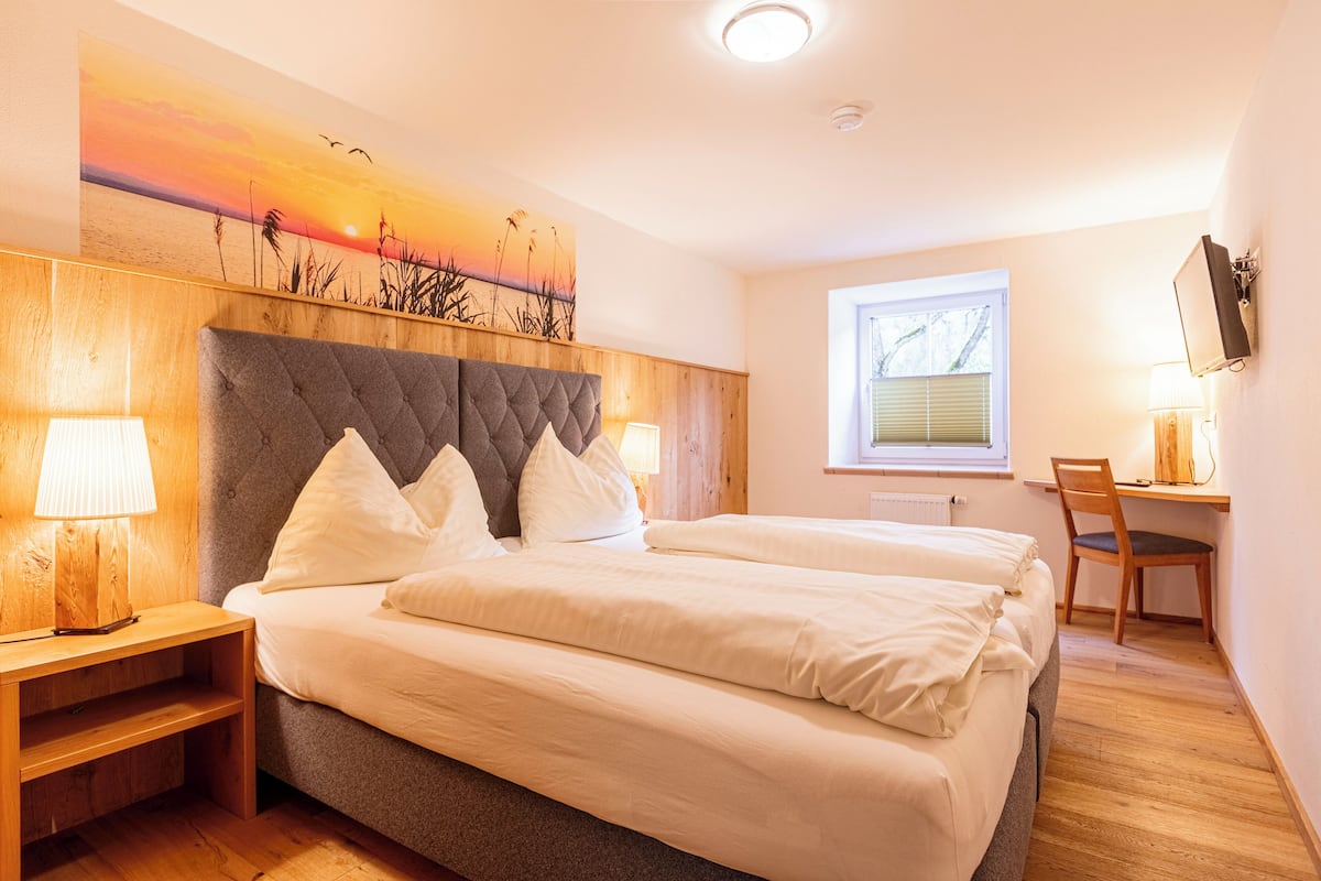 Quarto com cama grande, duas mesas de cabeceira com abajures, cabeceira de madeira, escrivaninha com cadeira e janela com persianas.