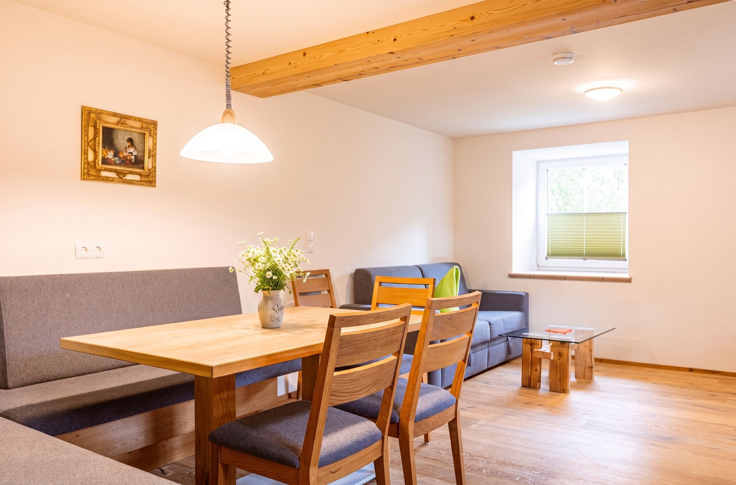 A dining area with a wooden table, chairs, a vase with flowers, a sofa, and a window with blinds.