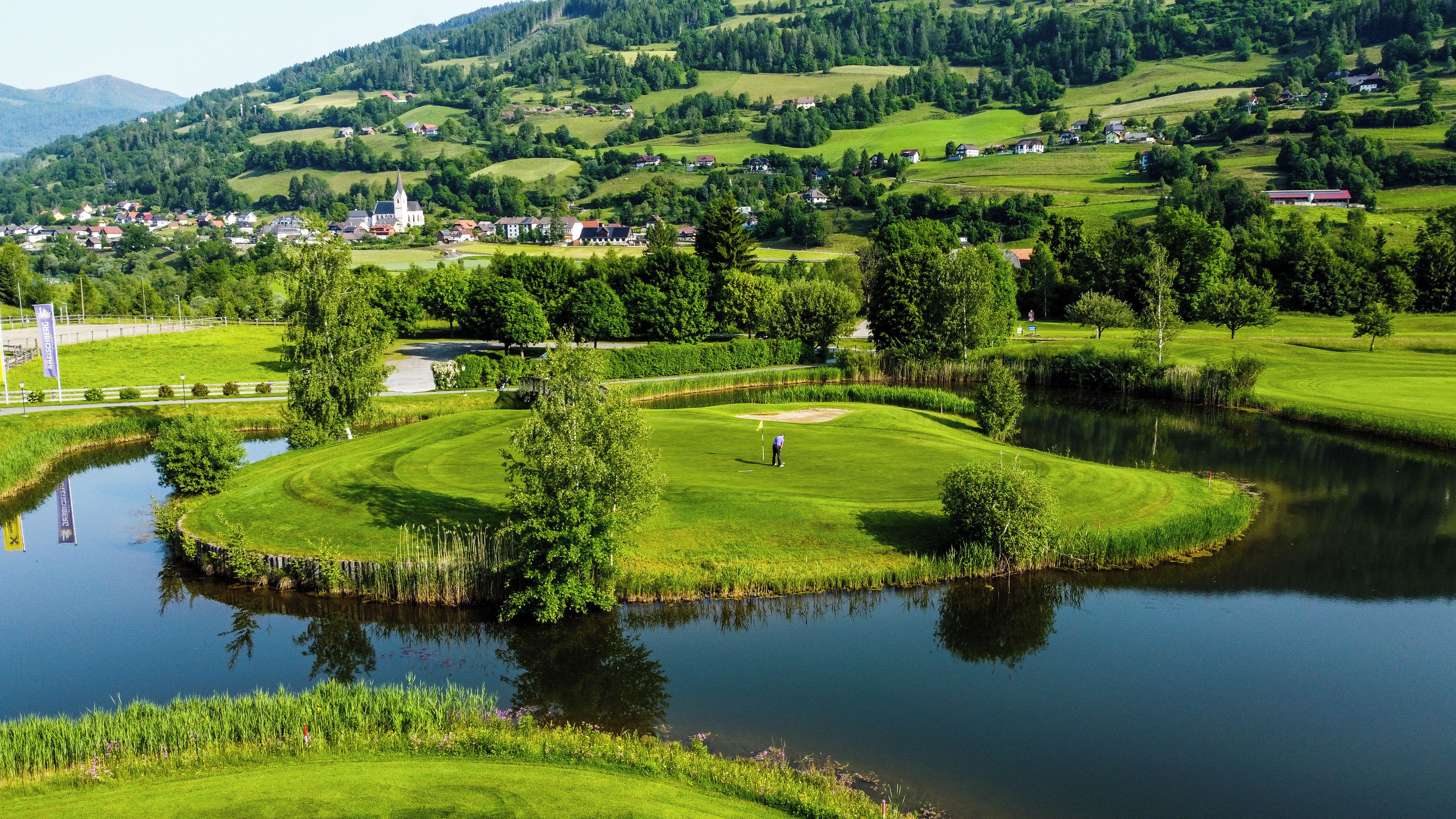 Um campo de golfe com um lago, rodeado por vegetação e uma pequena aldeia ao fundo.