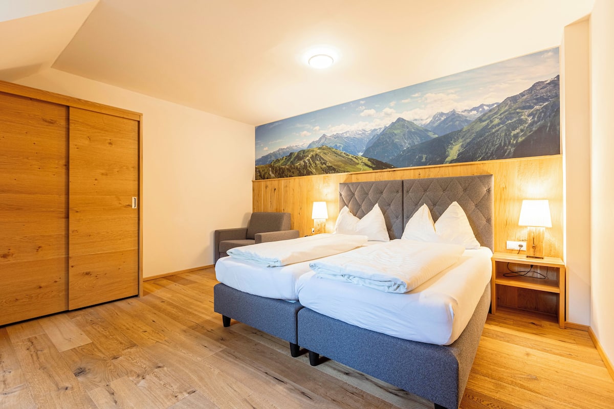 Uma cama de casal com cabeceira cinzenta, duas mesas de cabeceira com luminárias, um guarda-roupa de madeira e um mural na parede com paisagem de montanhas.