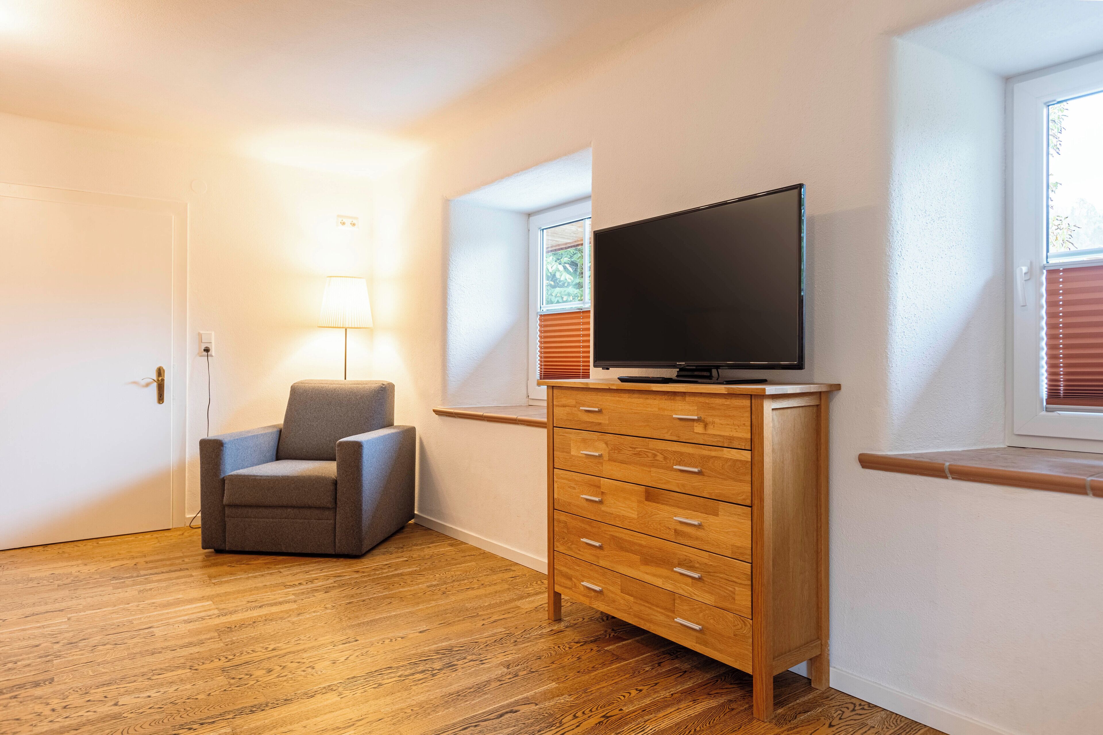 A room with a wooden floor, a grey armchair, a wooden dresser, a television on top, and a window with blinds.