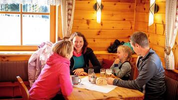 Interior de madeira com mesa de jantar, cadeiras e janela com cortinas de renda.